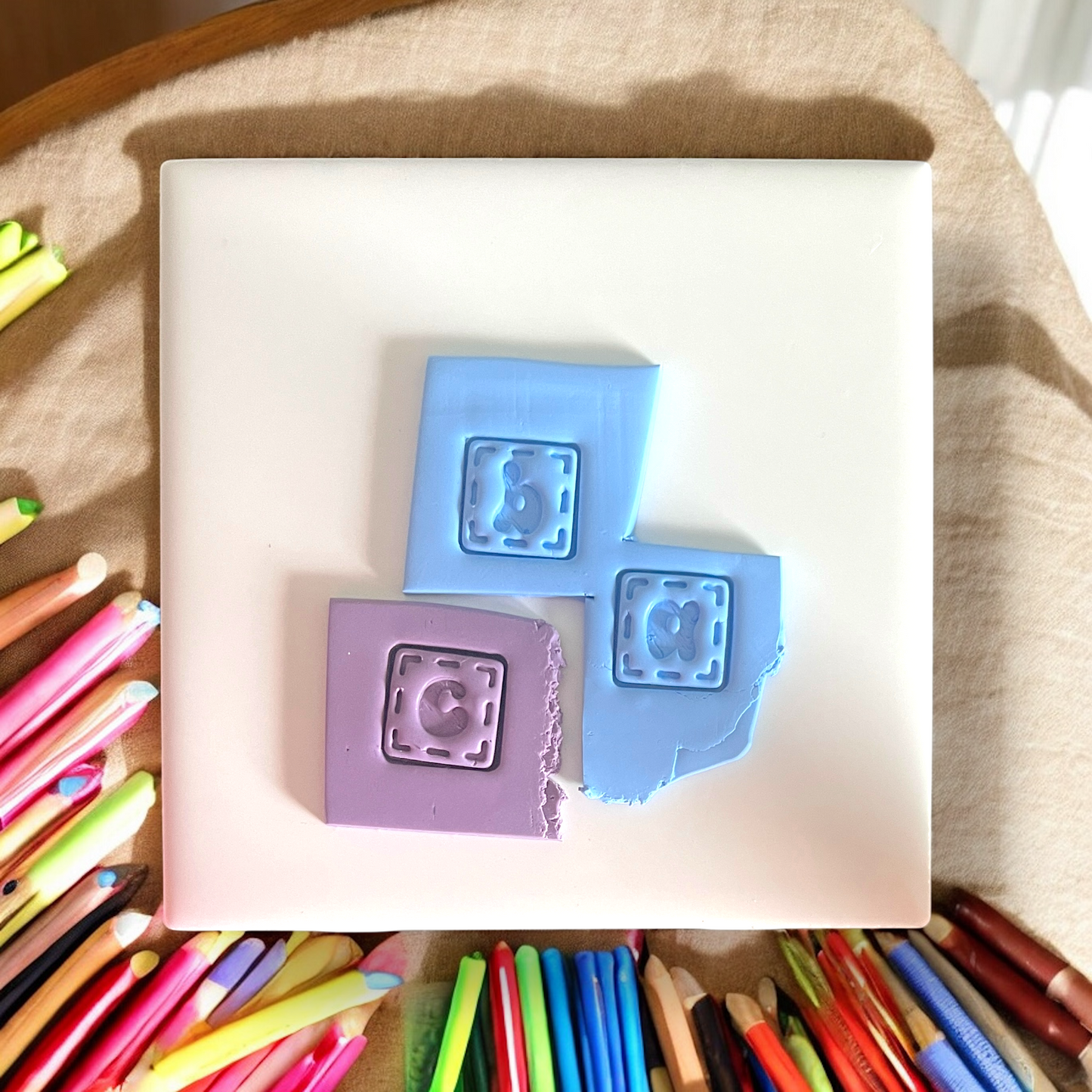 Two square silicone molds with letter stamps on a white surface, surrounded by colorful pens.