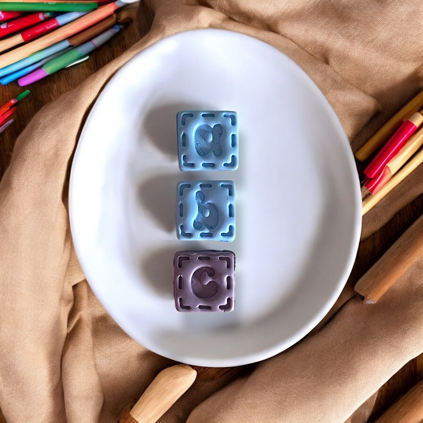 Two blue and one purple letter 'a' mold on a white plate with colored pencils and wooden sticks in the background.