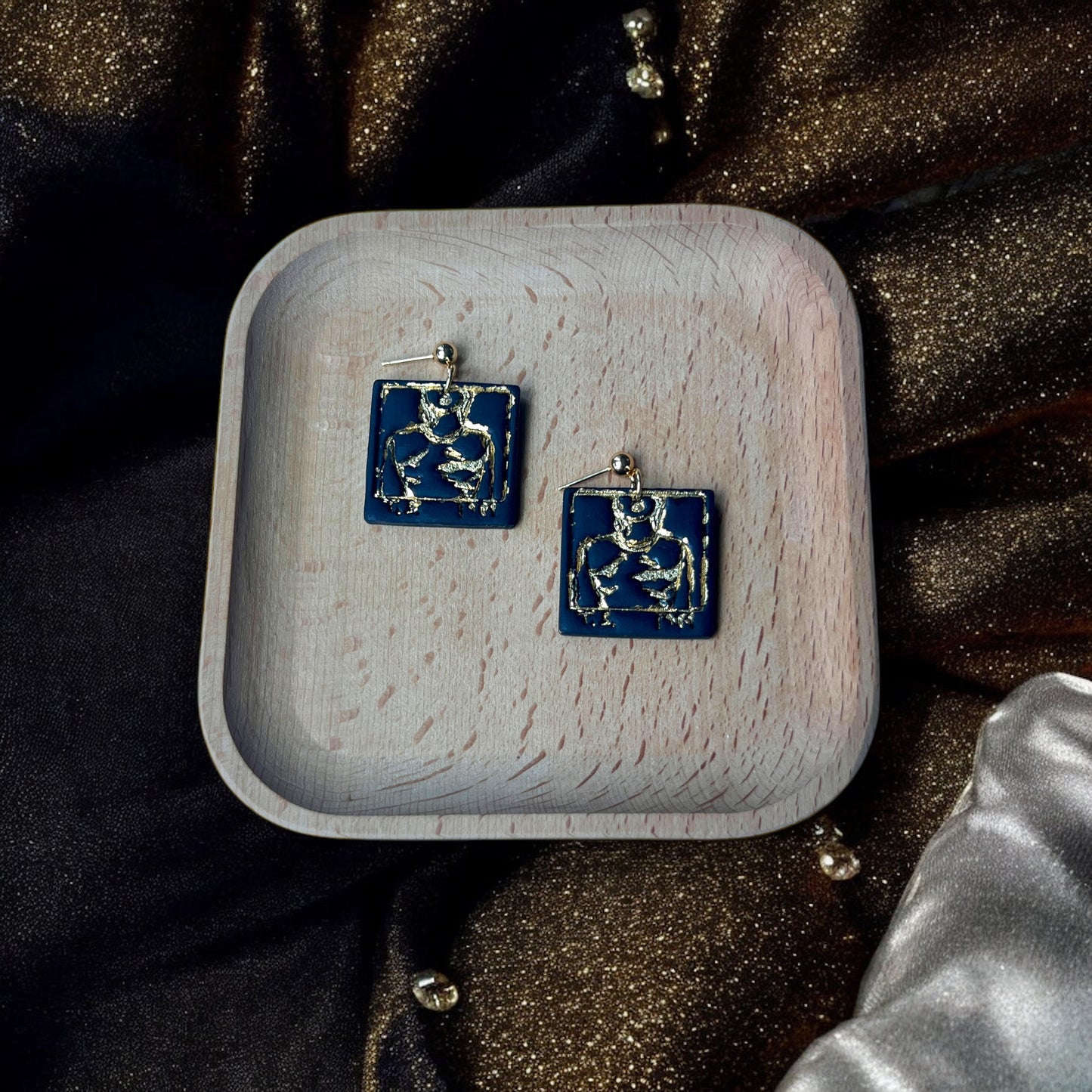 Square black earrings featuring abstract gold accents, displayed on a wooden tray with a luxurious setting of gold, black, and pearl decor.