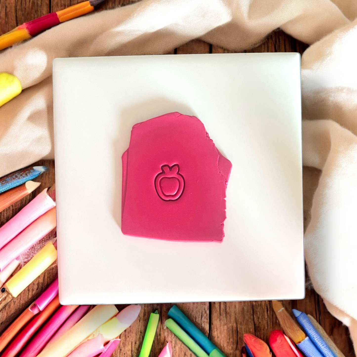 Apple-shaped clay impression on a dark pink slab surrounded by colorful pencils on a white ceramic tile.