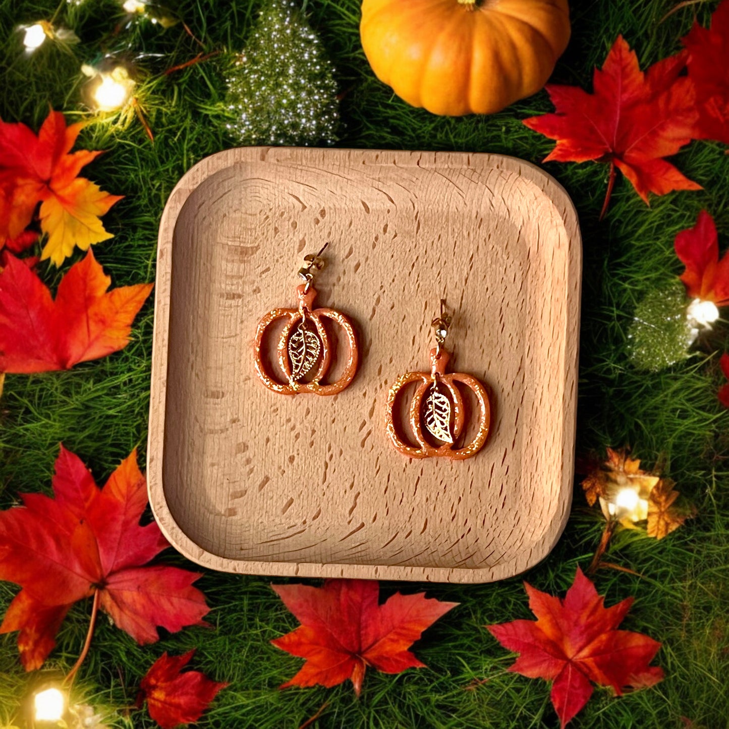 Pumpkin-shaped dangle earrings with glitter and gold leaf accents resting on a wooden tray, surrounded by vibrant fall leaves and glowing lights.