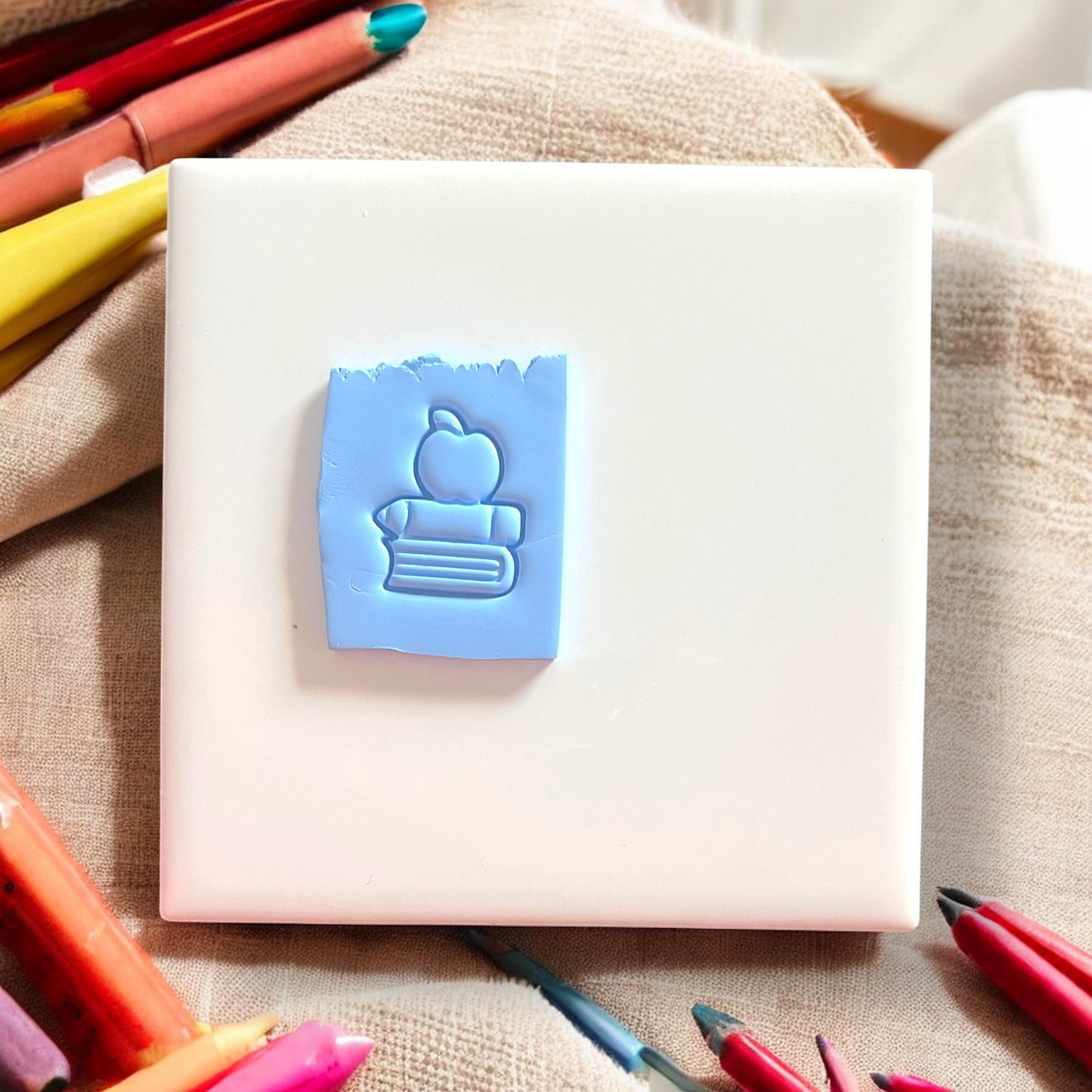White notebook with a blue rubber stamp featuring an apple and book design, surrounded by colorful markers on a textured surface.