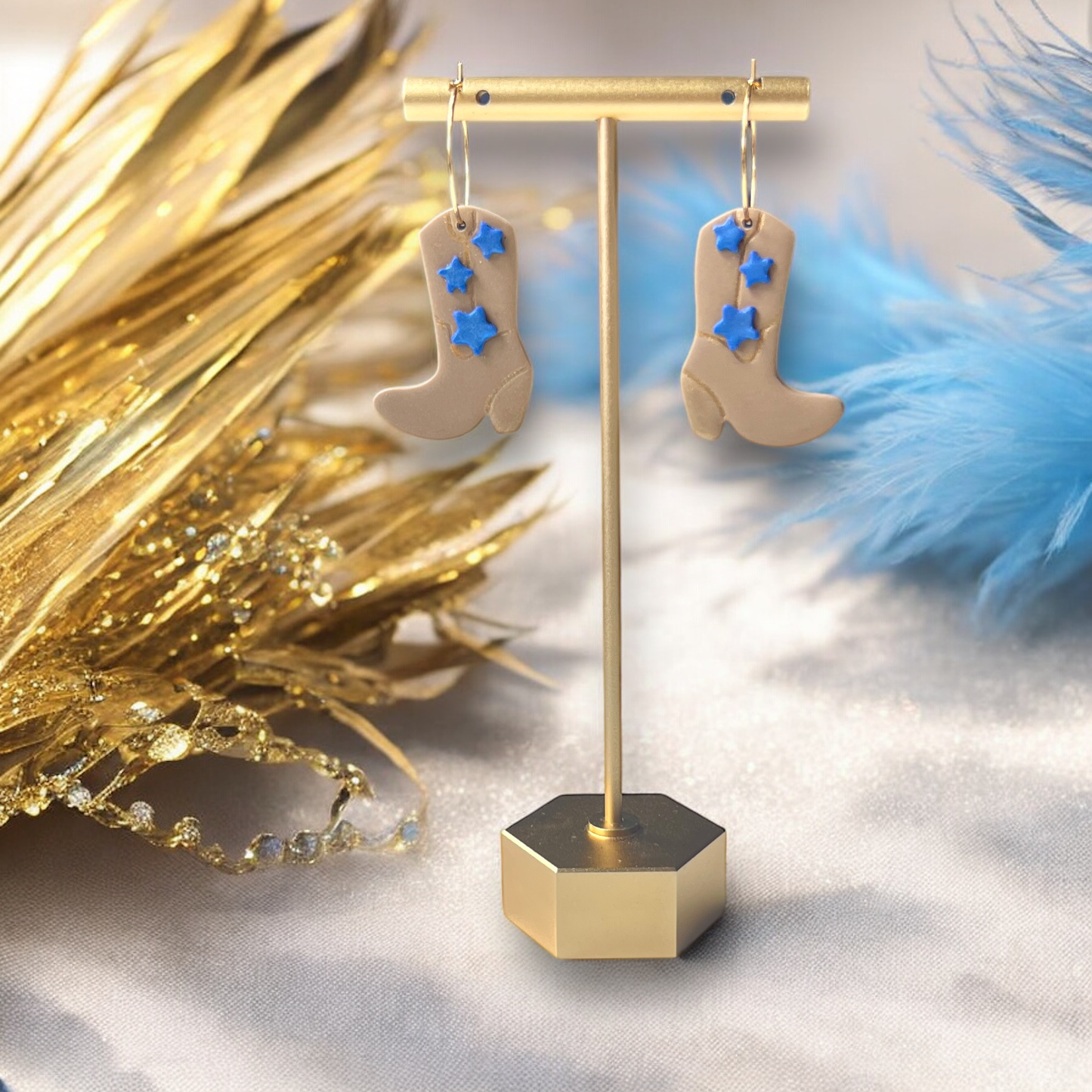 Beige boot-shaped earrings with blue star accents, hanging on a sleek gold stand, surrounded by shimmering gold and blue decor.