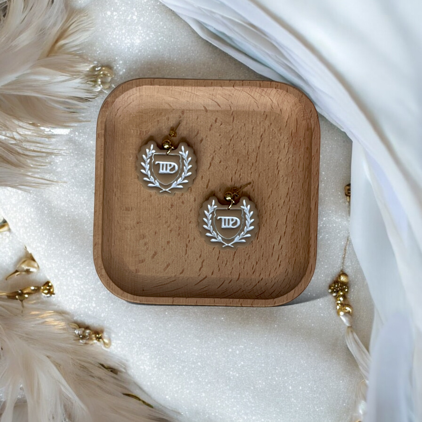 Beige laurel crest dangle earrings placed in a wooden tray, surrounded by soft white feathers and gold decorative elements.