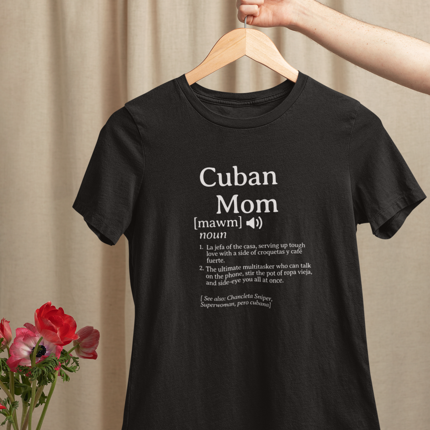Black Cuban Mom definition t-shirt hanging on a wooden hanger, displayed with nearby flowers against a soft neutral backdrop.