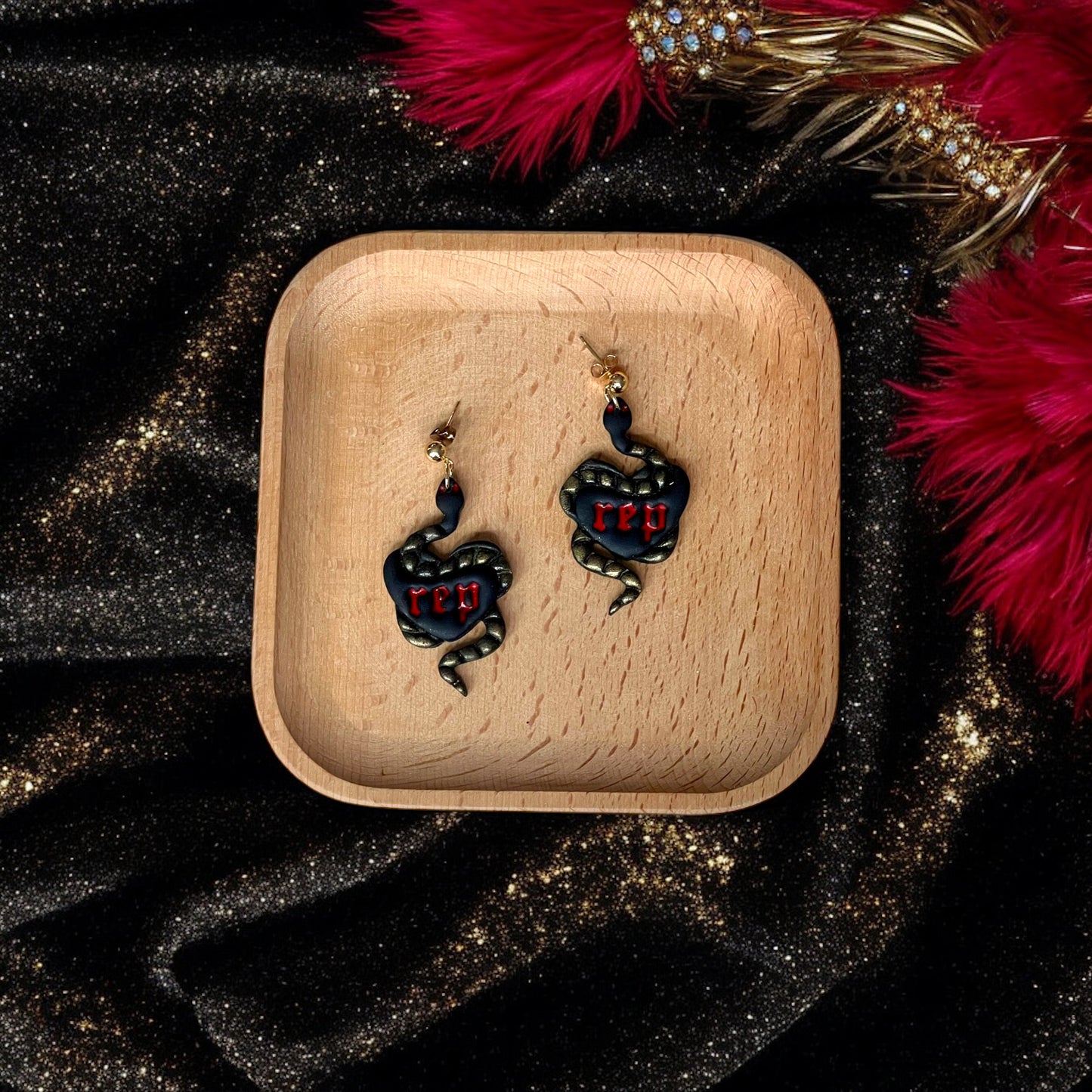 Pair of black heart earrings featuring coiled gold snakes and red “rep” text, resting on a wooden tray against a glittery black and red setting.