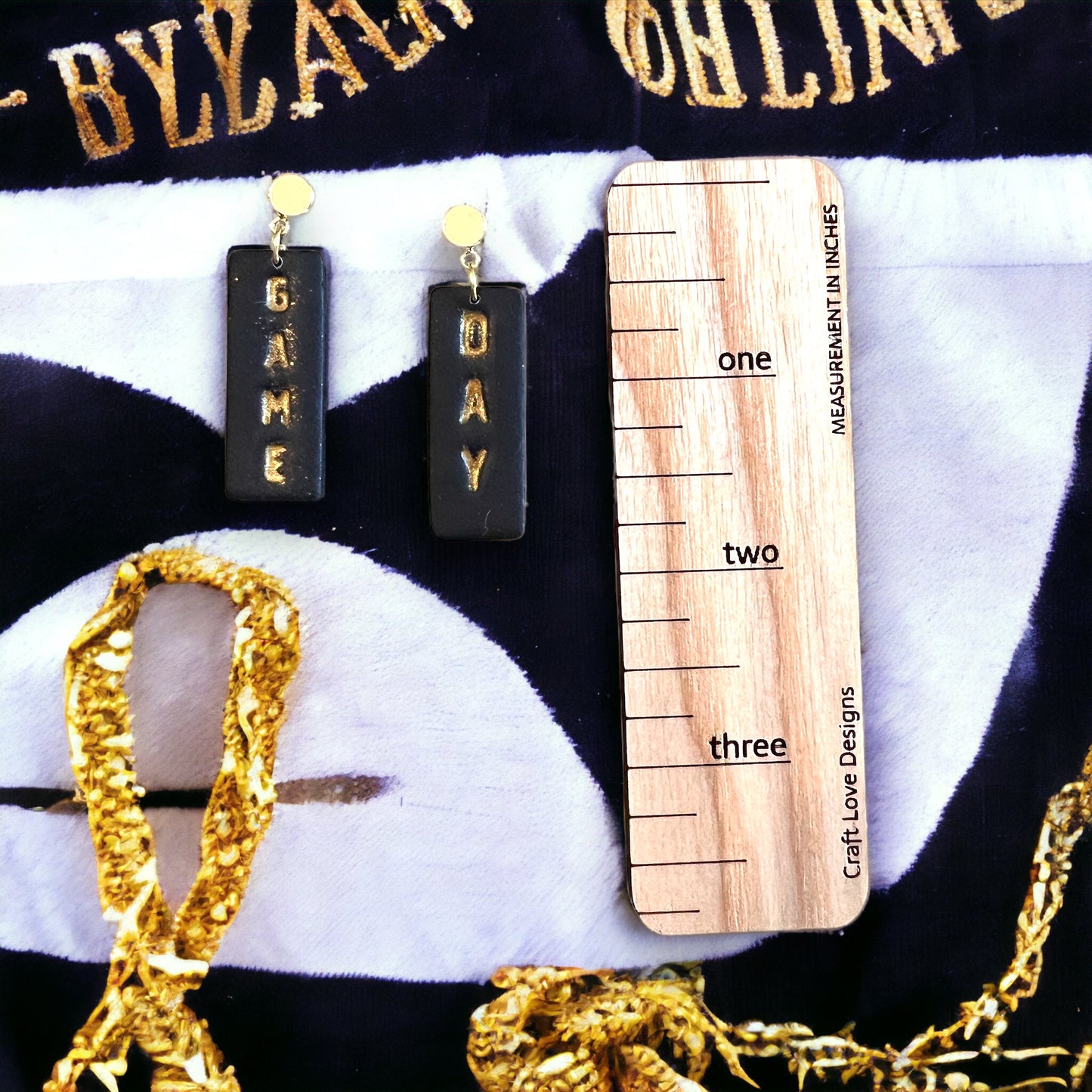 Black and gold customizable polymer clay earrings, showcasing bold "Game" and "Day" text, measured against a wooden ruler.