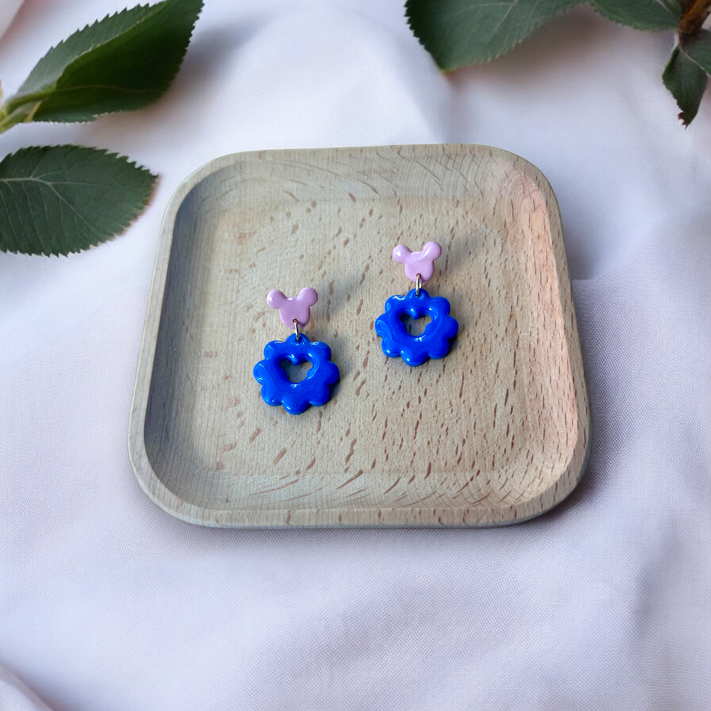 Blue flower earrings featuring a heart cutout and pink studs, neatly placed in a wooden tray on a fabric background surrounded by green leaves.