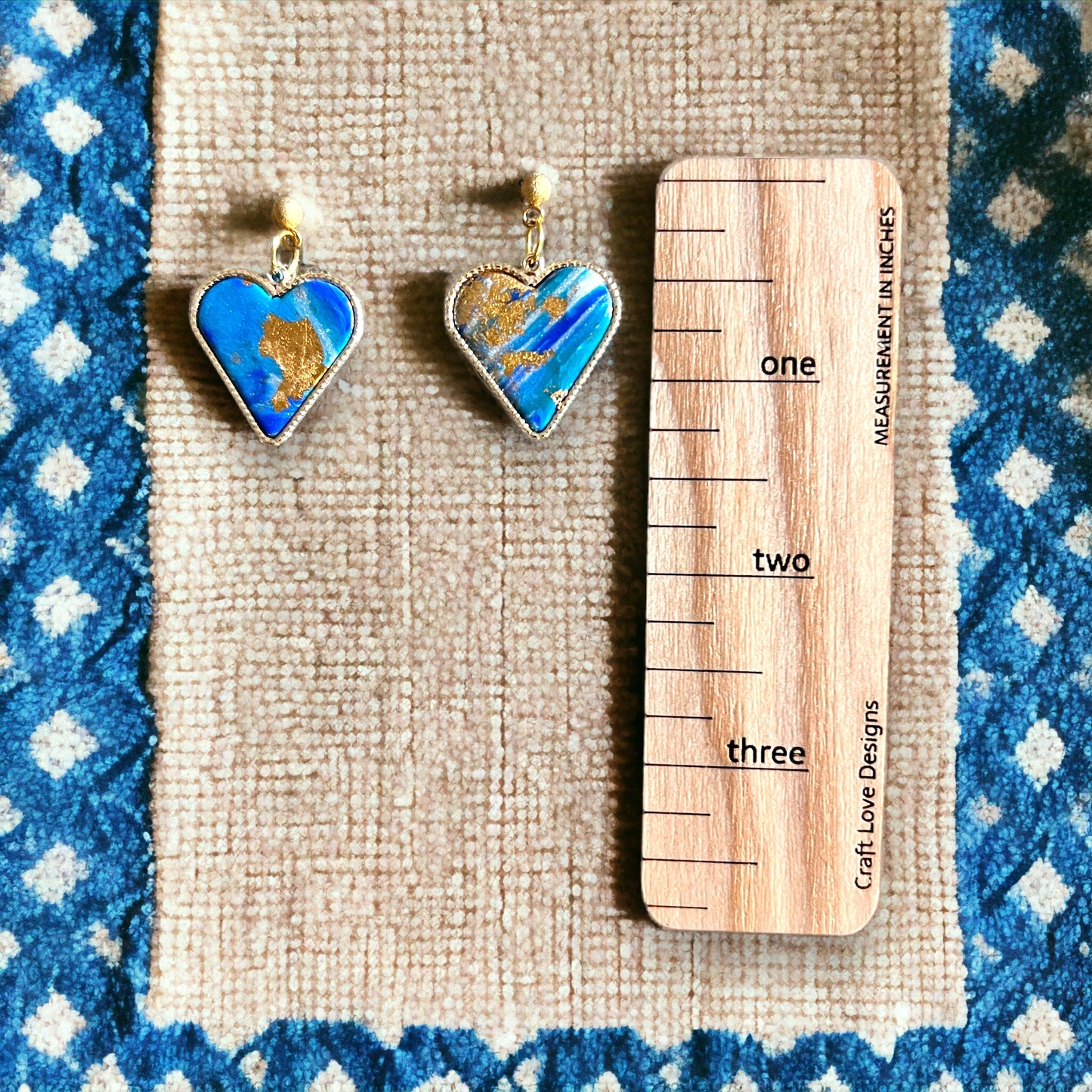 Blue and gold heart dangle earrings with a measuring ruler beside them, showcasing size comparison on a blue-patterned surface.