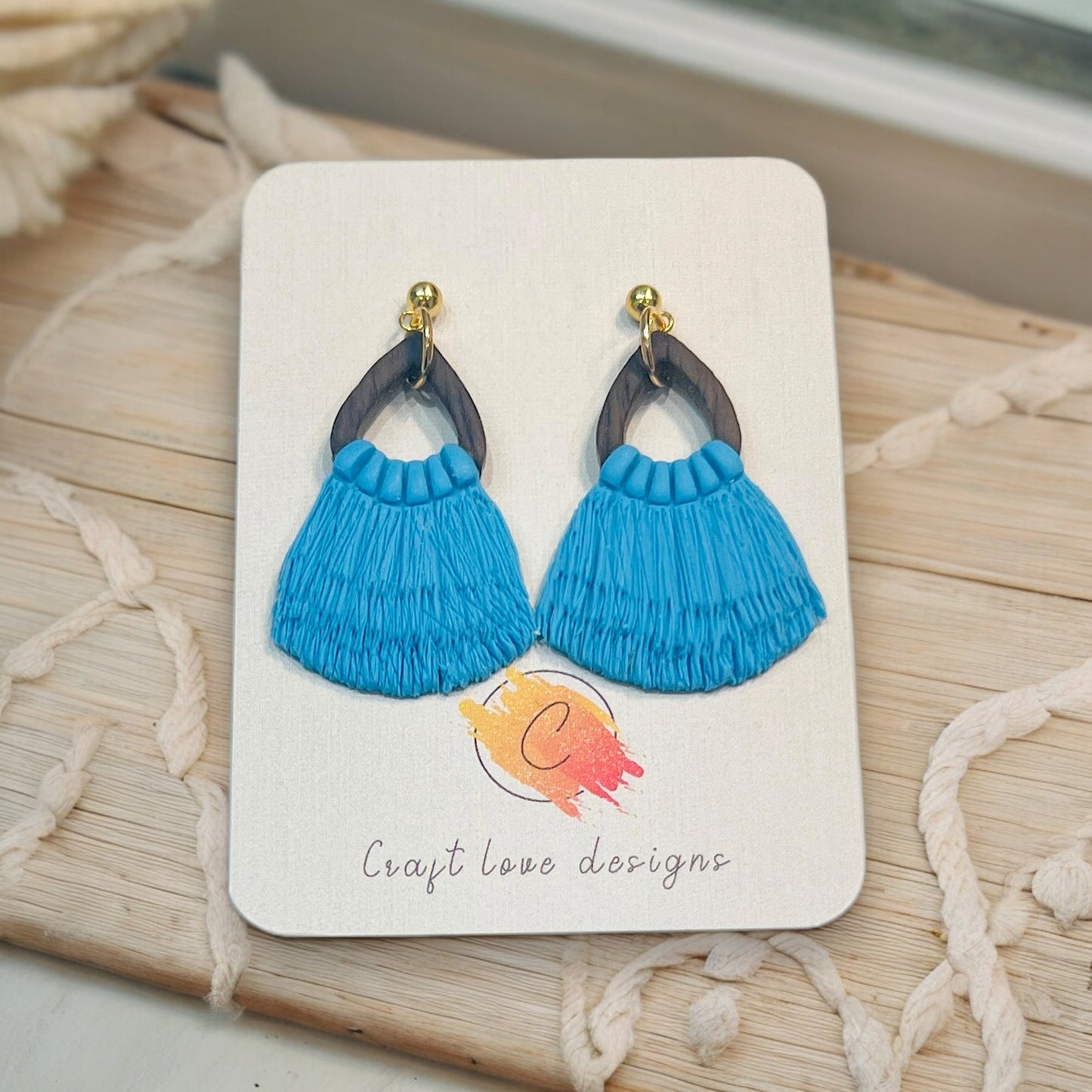 Blue macrame drop earrings with a wooden top frame and gold details, artistically displayed on Craft Love Designs' branded card.