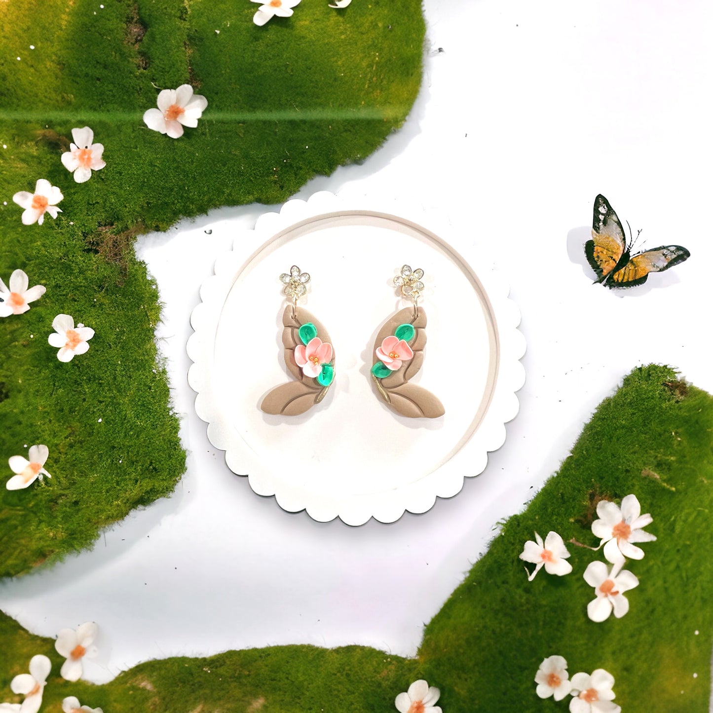 Latte and coral butterfly dangle earrings displayed among green moss, mini flowers, and a butterfly, creating a whimsical natural scene.
