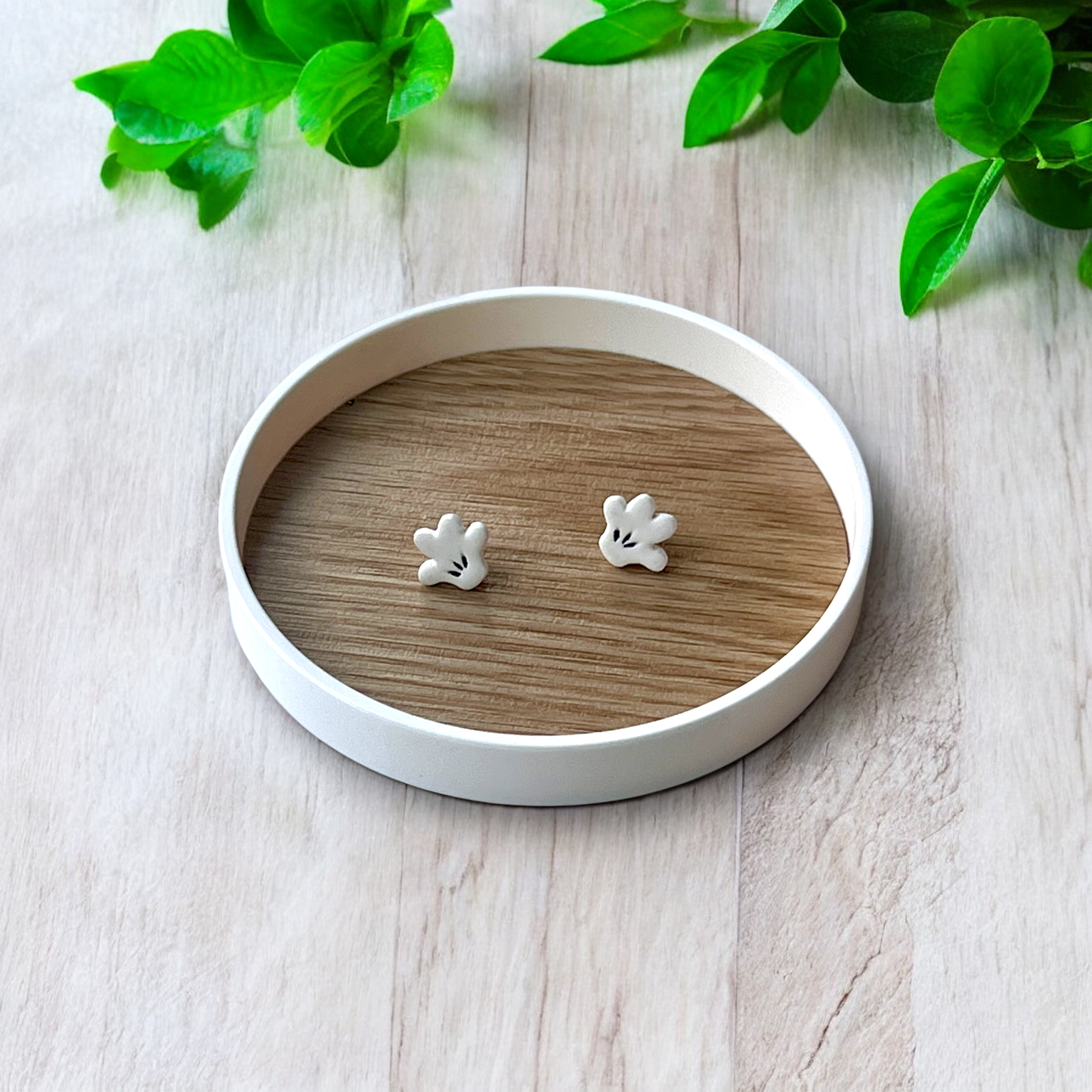 Cute white cartoon hand-shaped stud earrings displayed inside a round wooden tray placed on a light wood surface with greenery nearby.