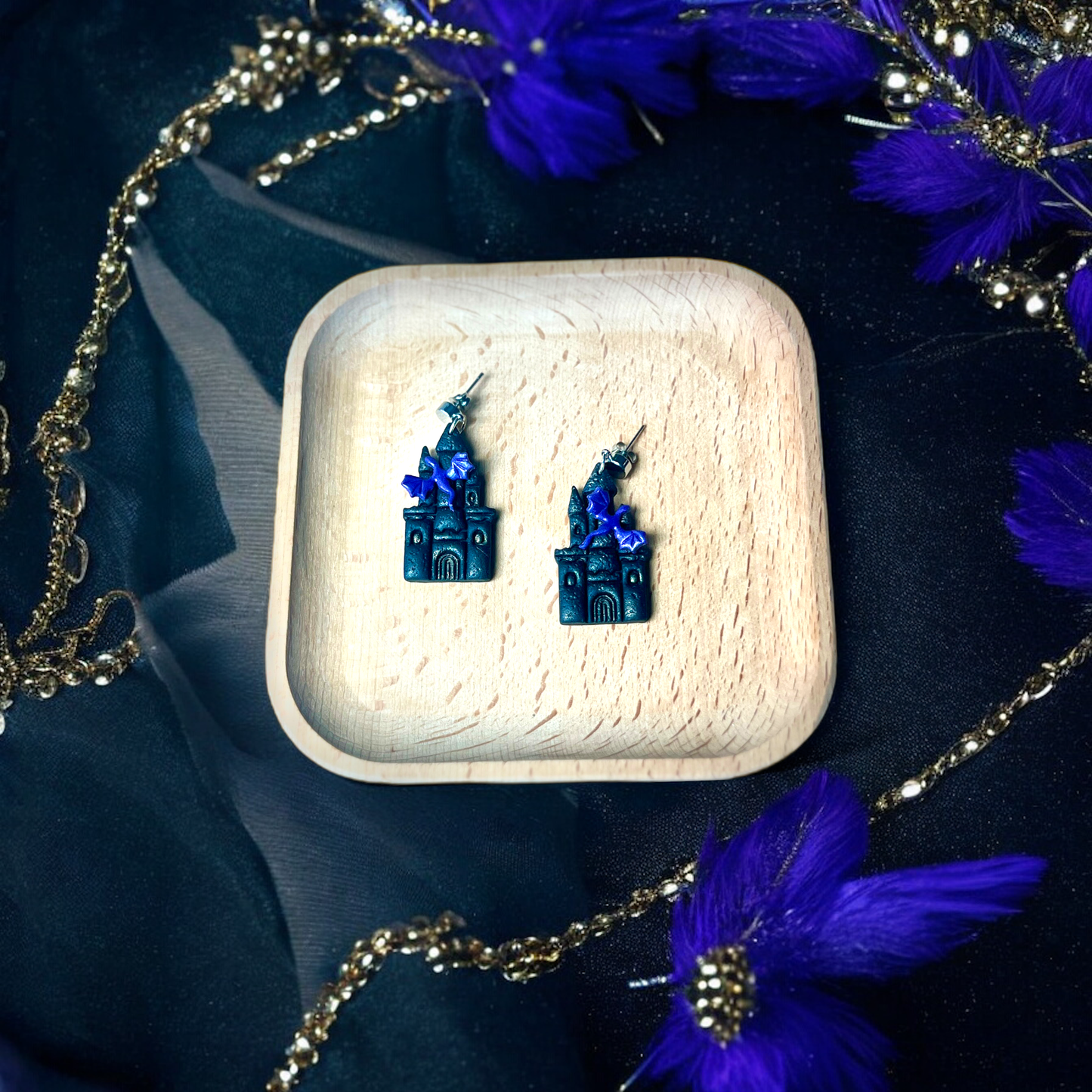 Castle earrings with purple bats resting in a wooden tray, surrounded by gold chains, purple feathers, and a dark mystical backdrop.