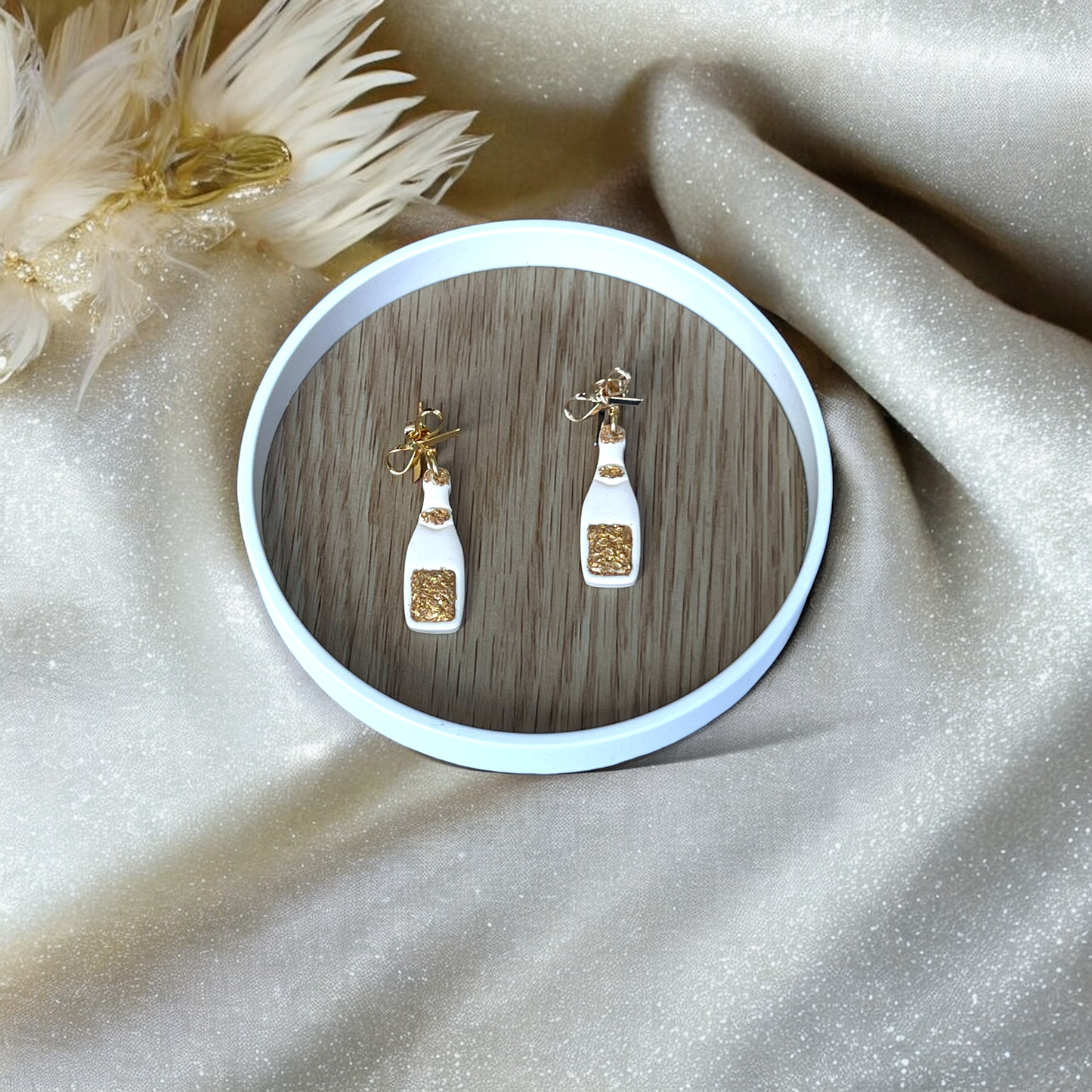 Elegant champagne bottle earrings with gold accents and bows resting in a round wooden tray, surrounded by soft, glittery fabric.