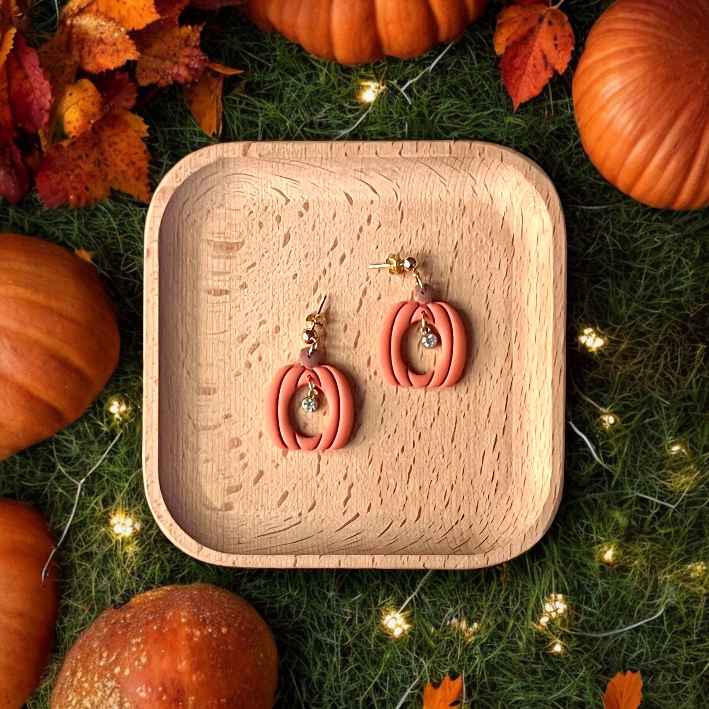 Festive clay pumpkin earrings featuring a crystal charm resting on a wooden tray, surrounded by fall pumpkins, leaves, and glowing fairy lights.
