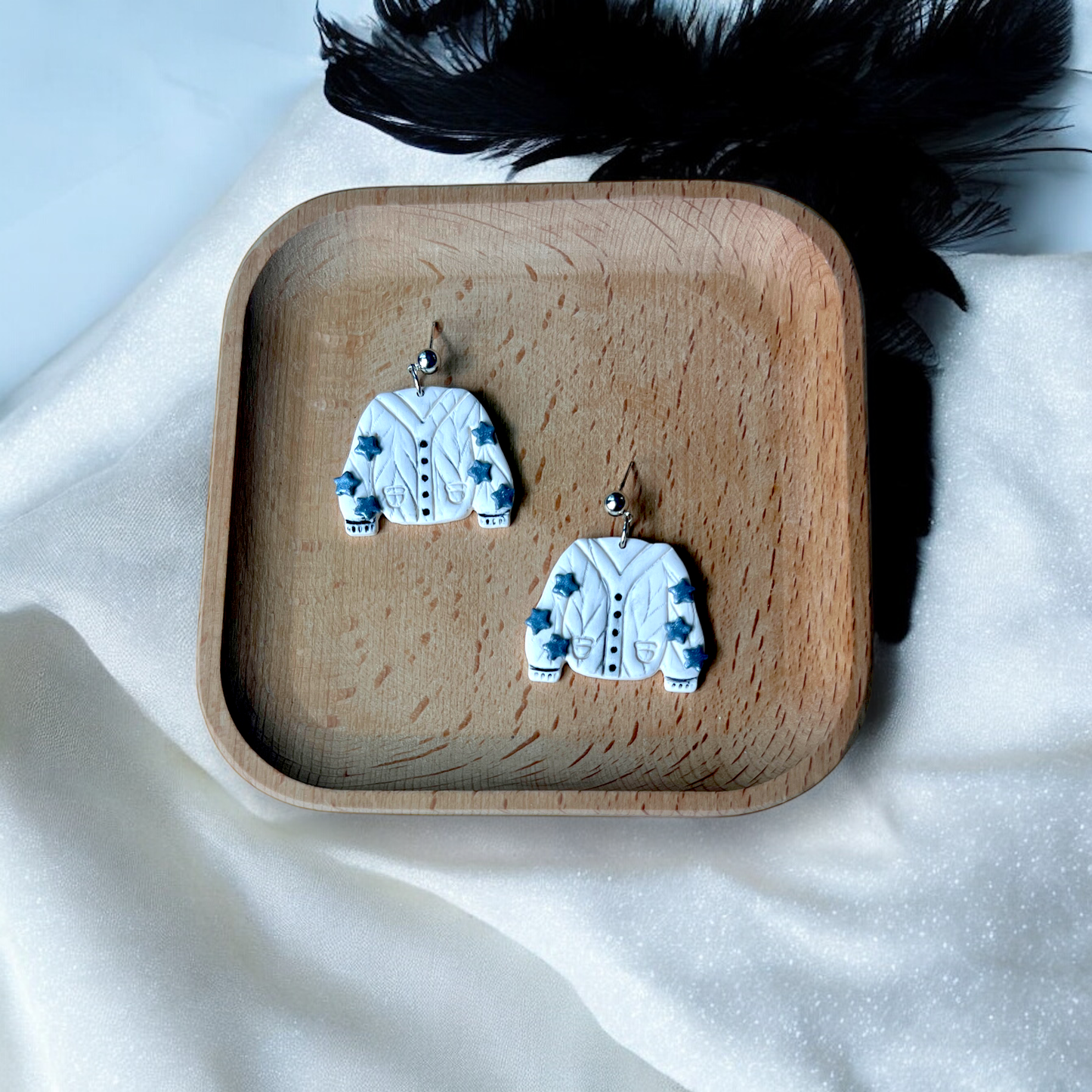 Cute cream sweater earrings with blue star details, displayed on a wooden tray with a soft beige fabric and black feather accents in the background.