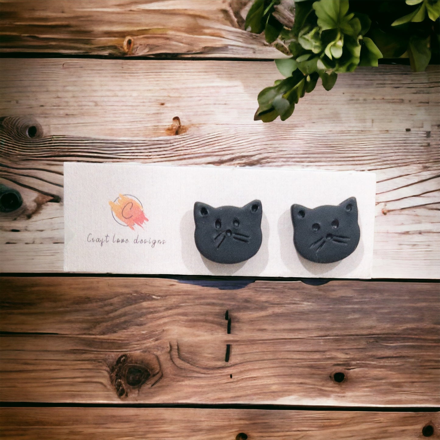 Black cat-shaped earring studs displayed on a Craft Love Designs card, placed on a rustic wooden background with green plants on the side.