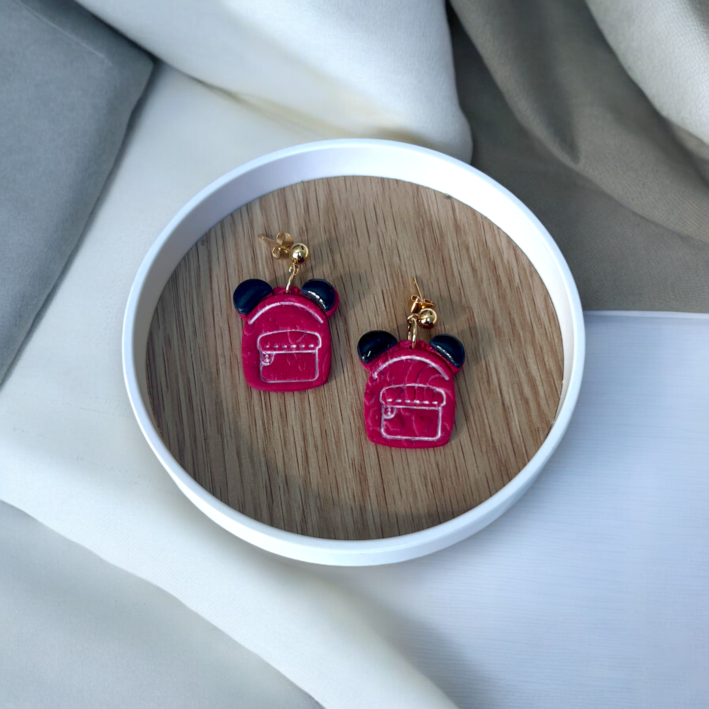 Adorable red backpack-shaped earrings with black ear details and gold studs, placed in a round wooden dish on neutral-colored fabrics.