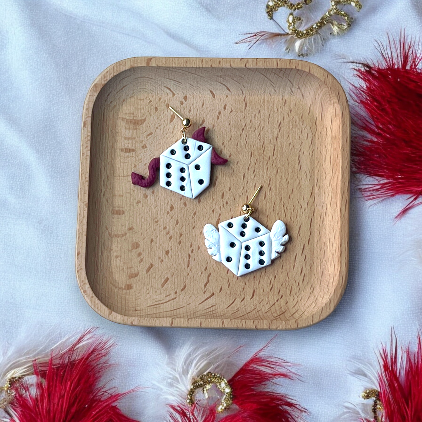 Pair of asymmetric dangle earrings with devil-horned dice and angel-winged dice, placed on a wooden tray surrounded by festive red feathers.