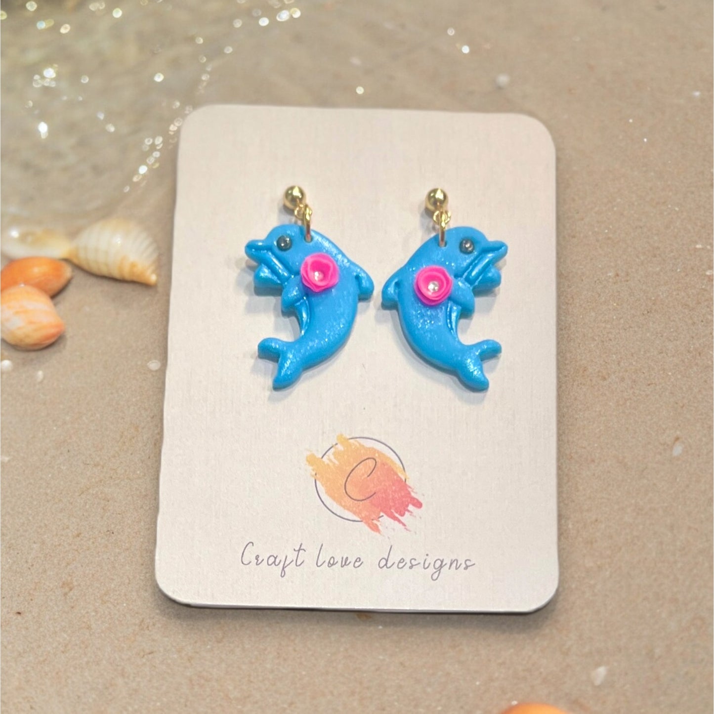 Handcrafted blue dolphin earrings with pink accents, presented on a Craft Love Designs display card, set on a sandy background near seashells.