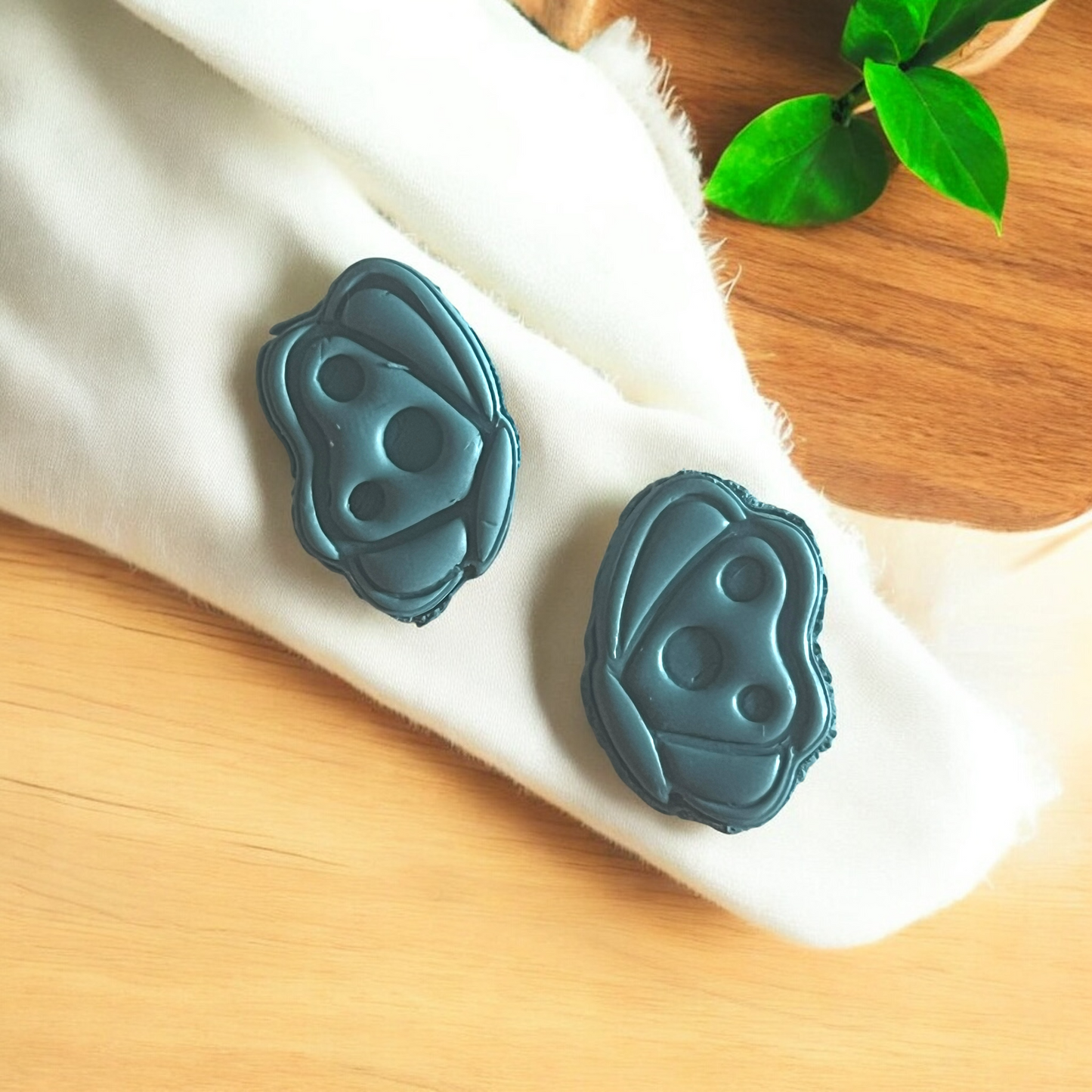 Teal earrings on a white cloth with a wooden surface and green leaves in the background