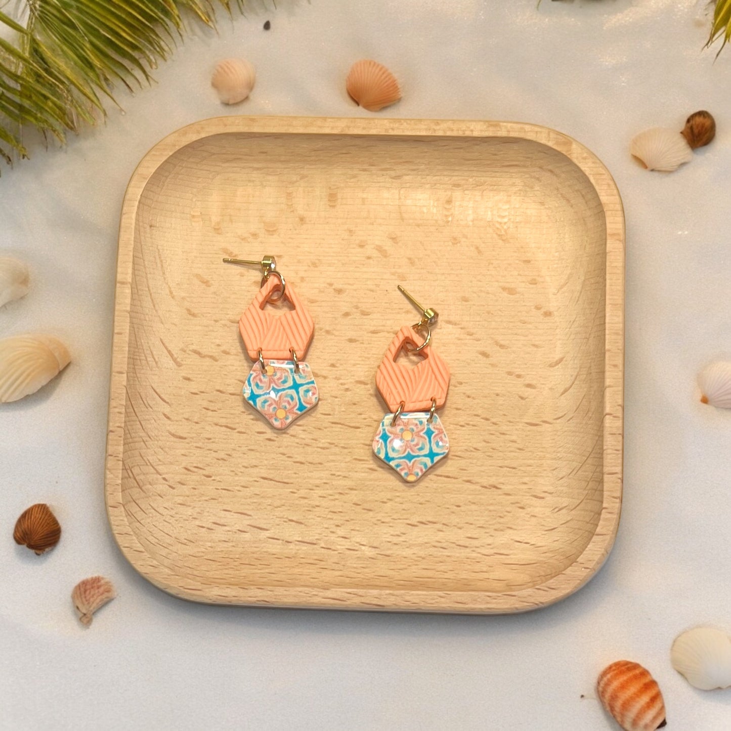 Chic drop earrings with orange textured tops and patterned bottoms displayed on a wooden tray amid scattered seashells