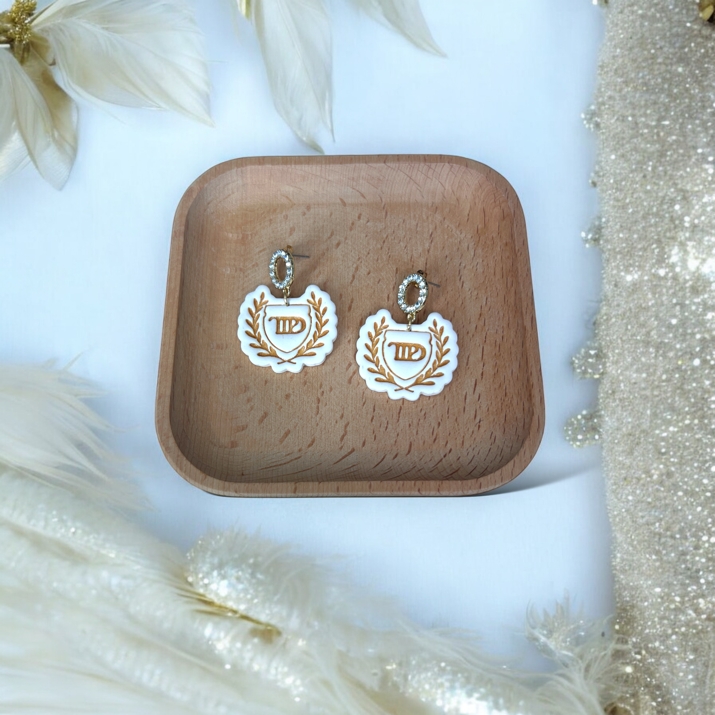 Elegant white laurel crest earrings with gold detailing and rhinestone studs, placed in a wooden tray, surrounded by feathers and pearls.