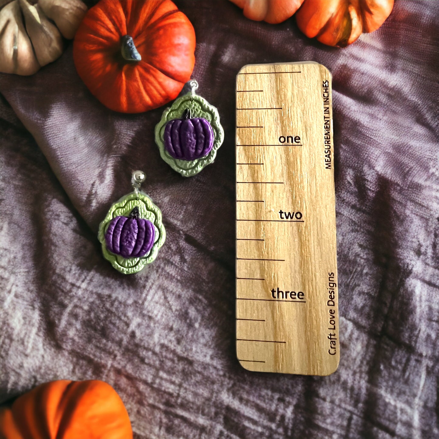 Close-up of handcrafted green and purple pumpkin earrings on a measurement tool, surrounded by autumn-themed pumpkins. Ideal for fall outfits.