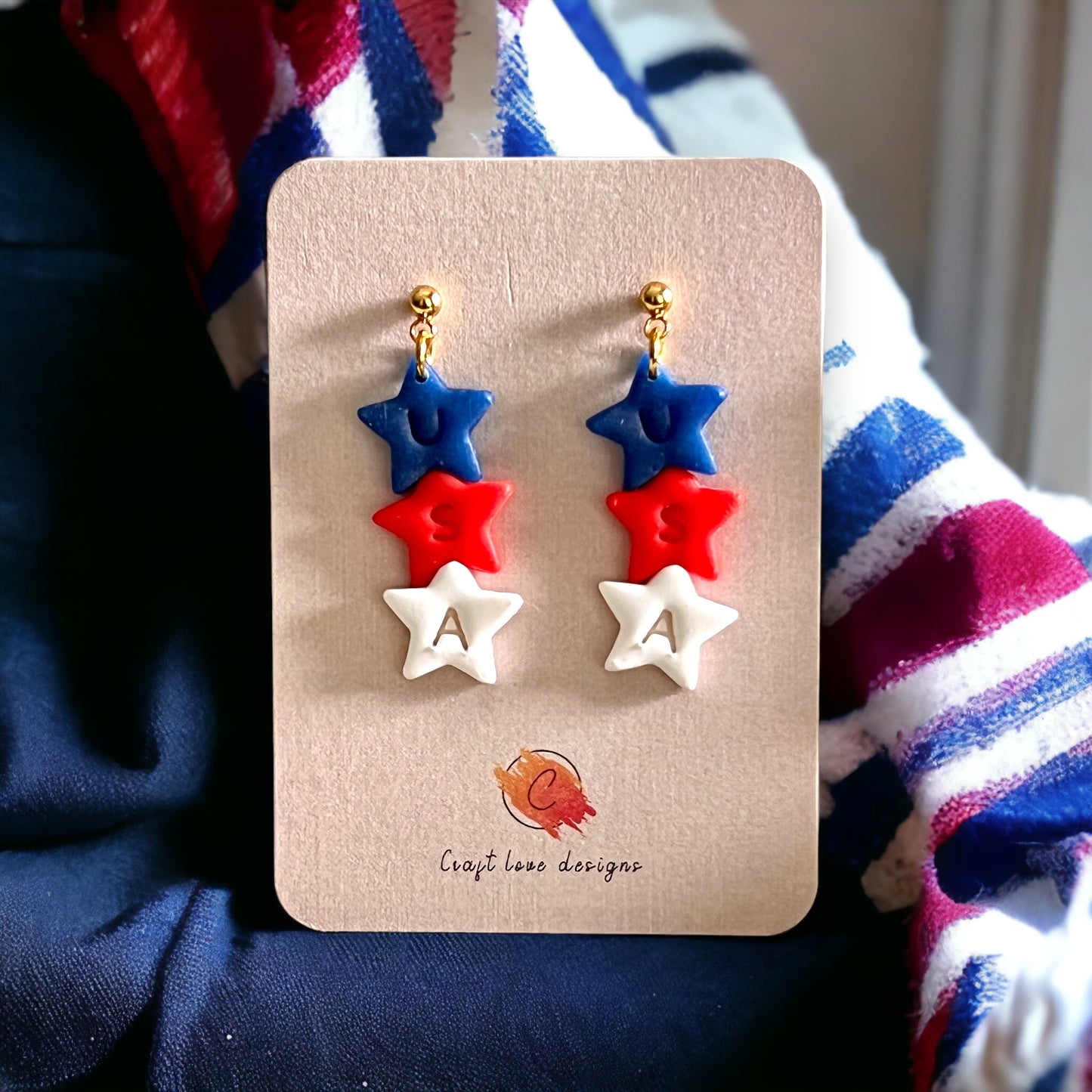 Patriotic USA-themed dangle earrings featuring red, white, and blue stars with gold hardware, on an earring card.