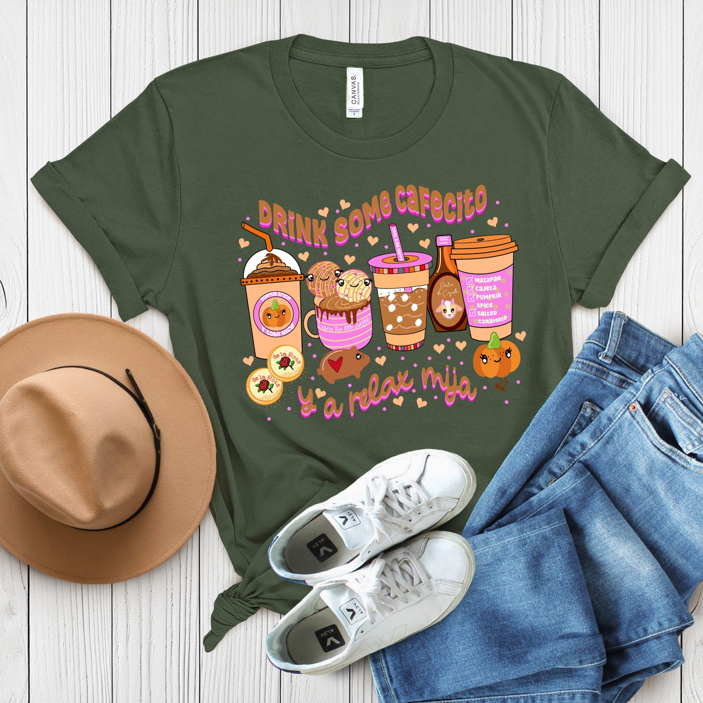 Flat lay of green "Drink Some Cafecito y a Relax Mija" t-shirt with coffee cup graphics, paired with a tan hat and sneakers on wood surface.