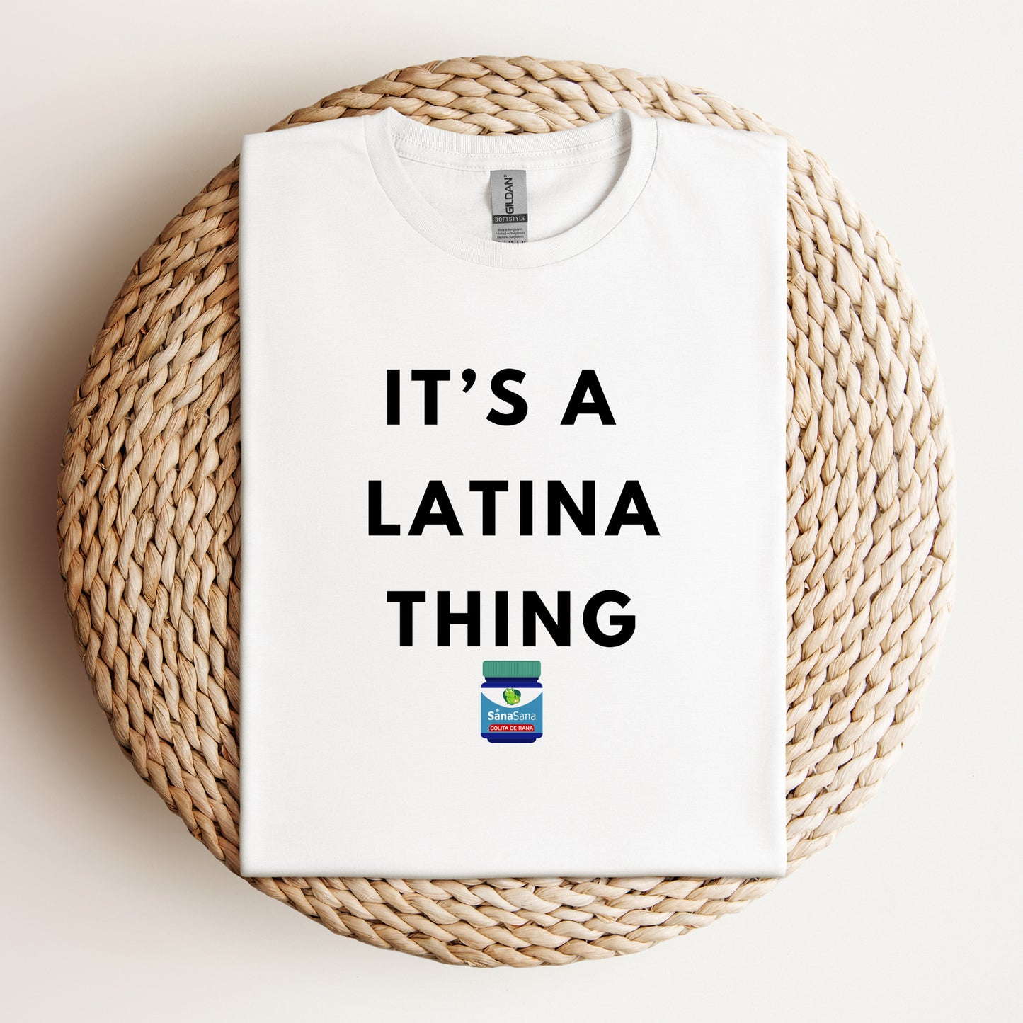 Flat lay of a white t-shirt with bold black text reading "It's a Latina Thing" and a small Vicks-inspired design, displayed on a woven background.
