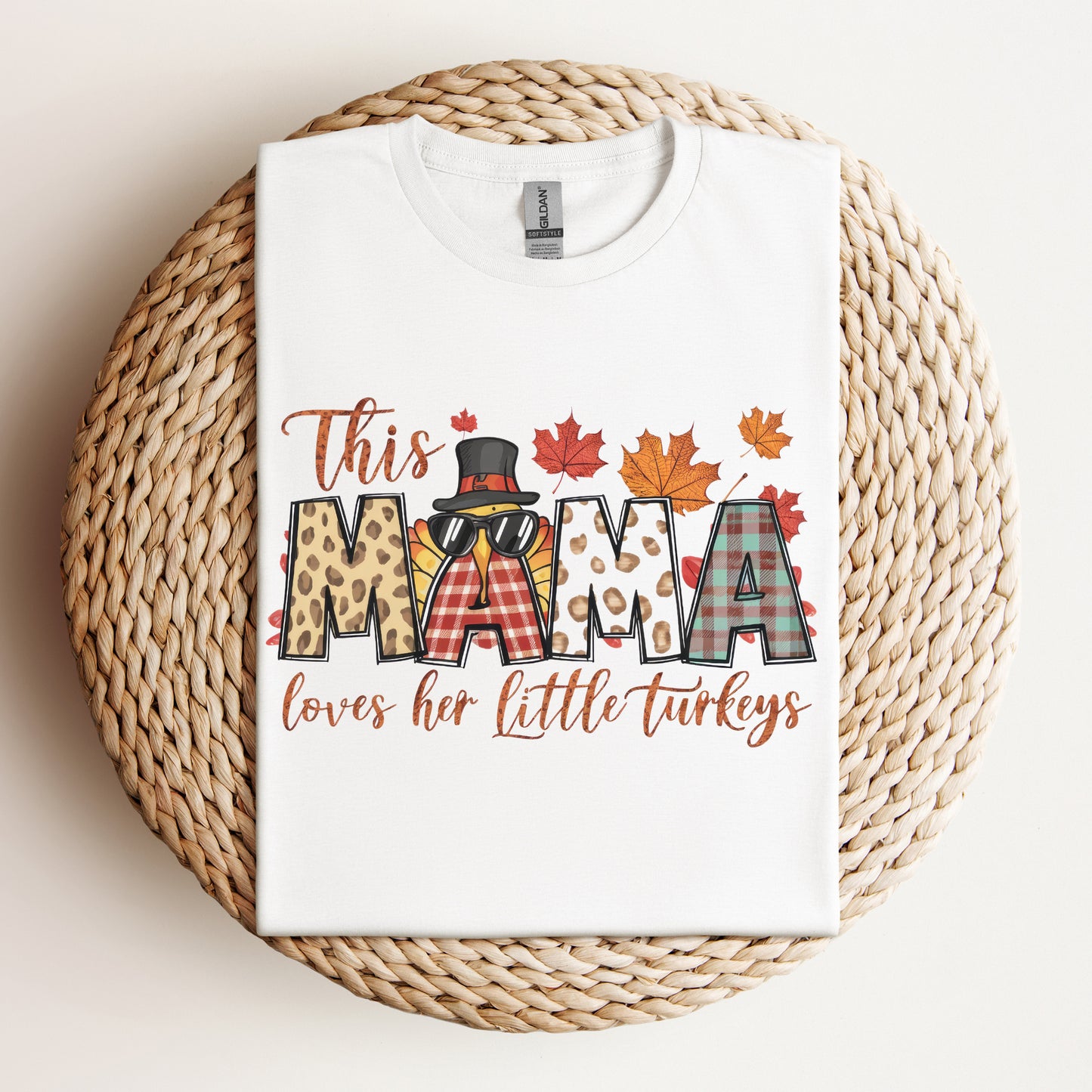 Flat lay of a white Thanksgiving-themed t-shirt with the text "This Mama Loves Her Little Turkeys" featuring fall leaves, plaid and leopard.