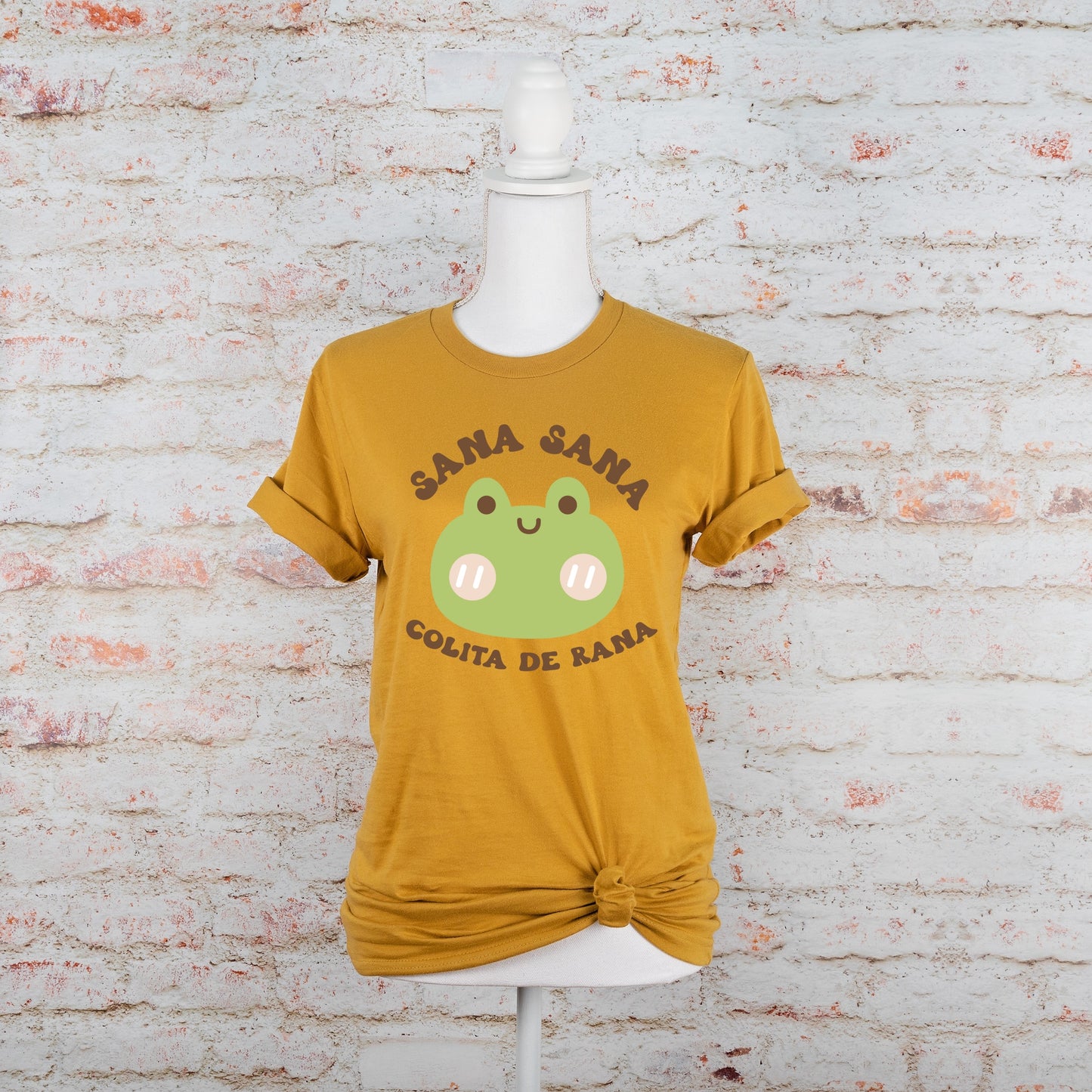 Flat lay of mustard yellow t-shirt with a green frog and Spanish phrase "Sana Sana Colita de Rana," placed on a neutral brick background.