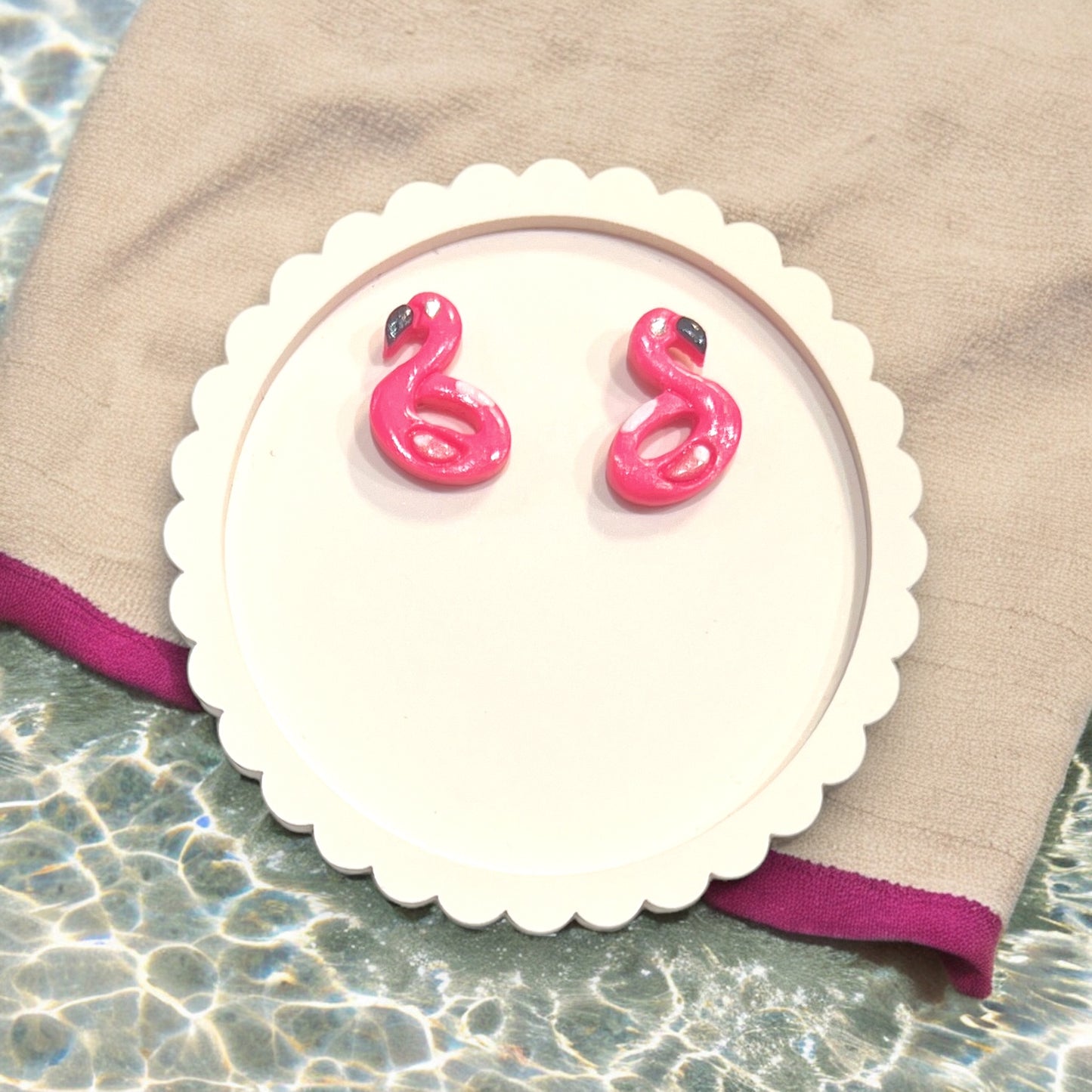 Pink flamingo earrings resting on a round decorative plate with a beach-themed backdrop. Perfect playful jewelry for summer outings.