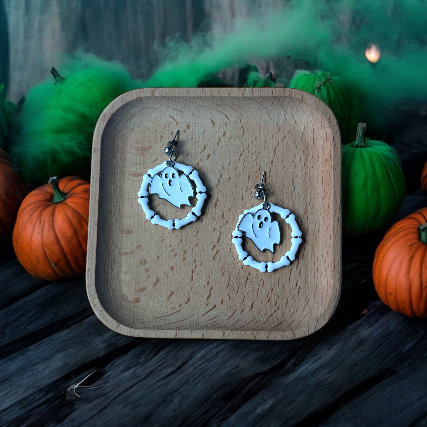 Ghost and bone hoop earrings resting on a wooden tray, surrounded by pumpkins and eerie green fog for a spooky Halloween theme.