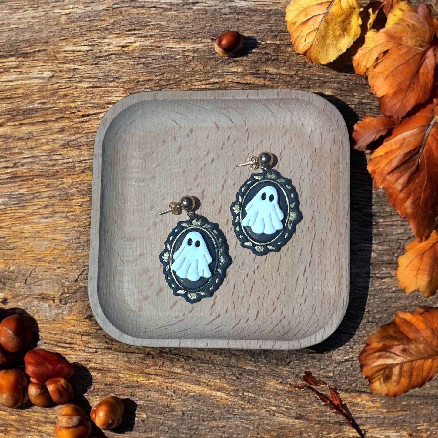 White ghost cameo earrings with black and gold frames resting in a wooden tray, surrounded by autumn leaves and warm seasonal decor.