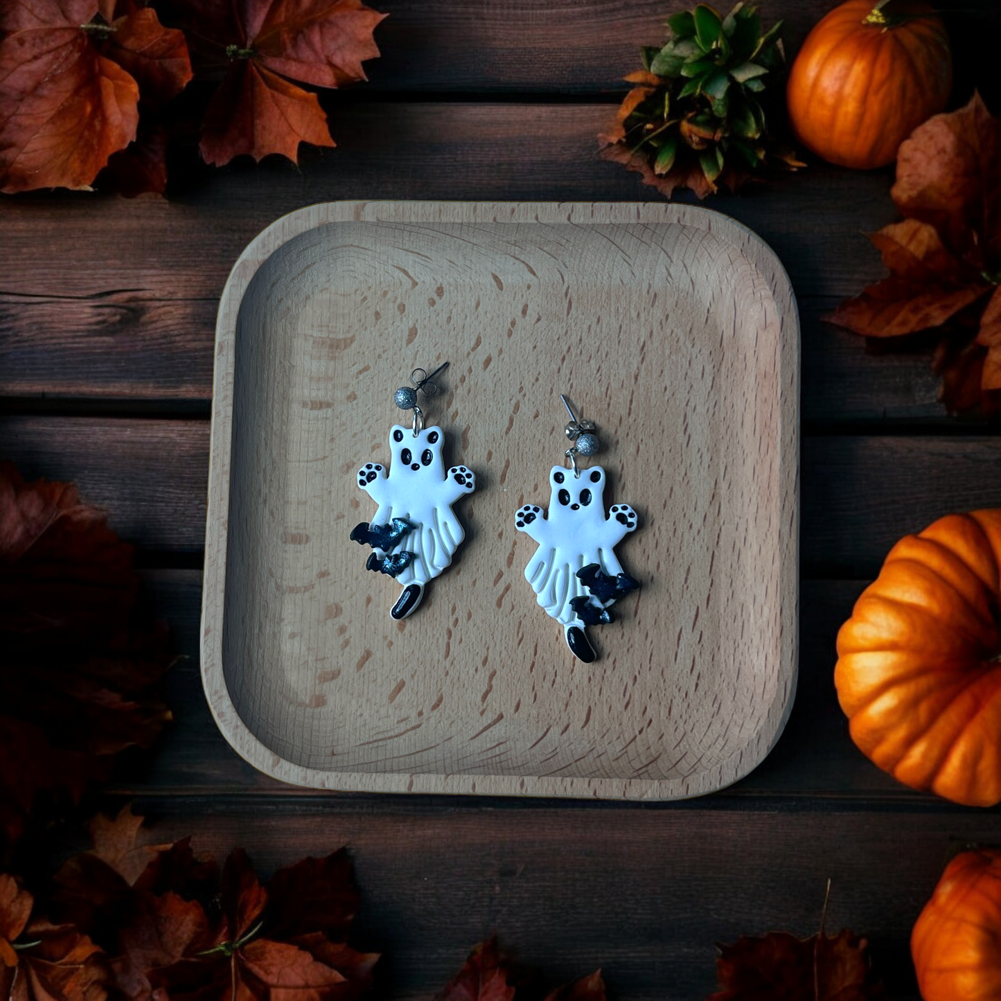 White ghost earrings with paw prints and black bat accents resting in a wooden tray, surrounded by pumpkins and fall leaves.