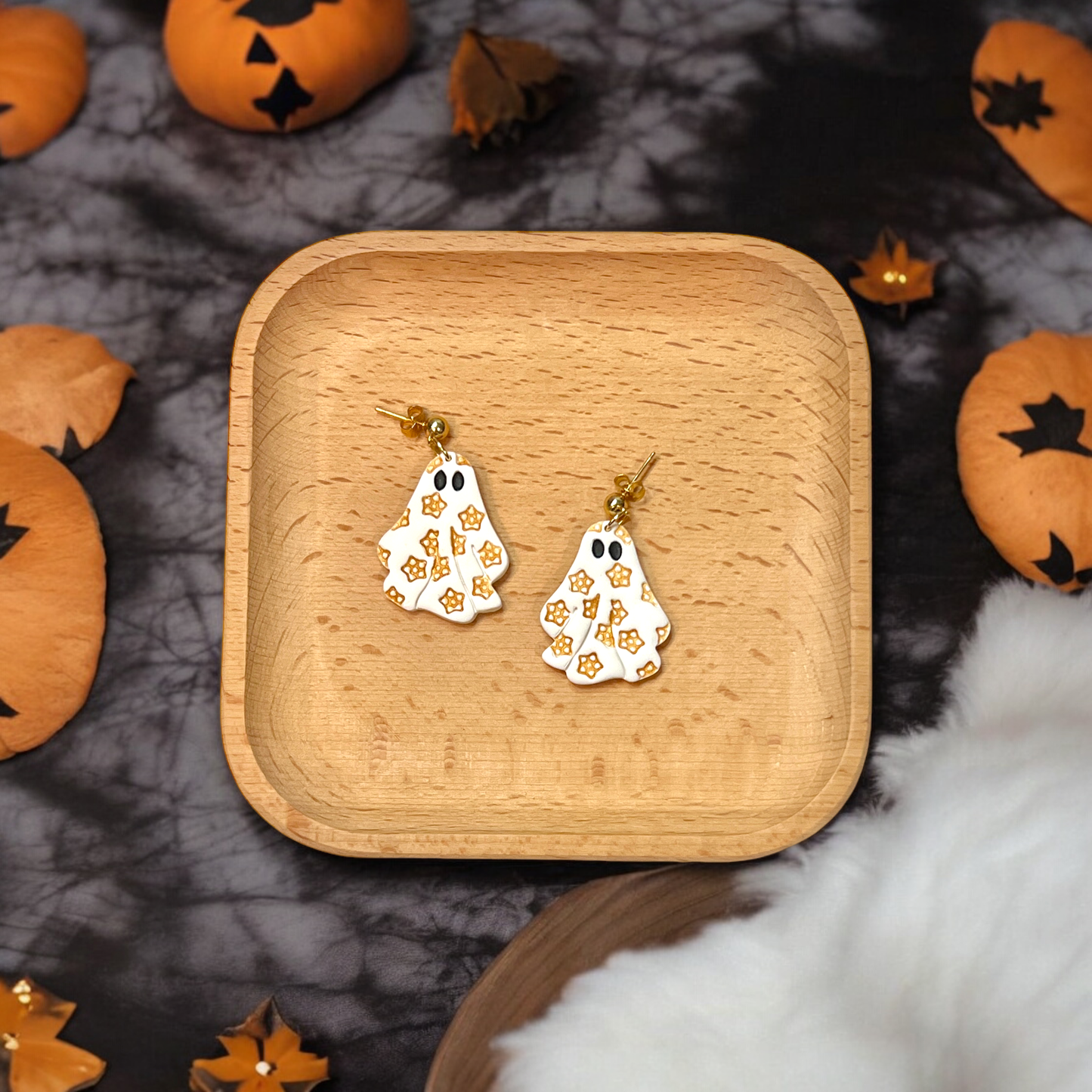 A close-up of small polymer clay ghost earrings decorated with gold flowers, presented on a wooden tray surrounded by Halloween-themed decorations.