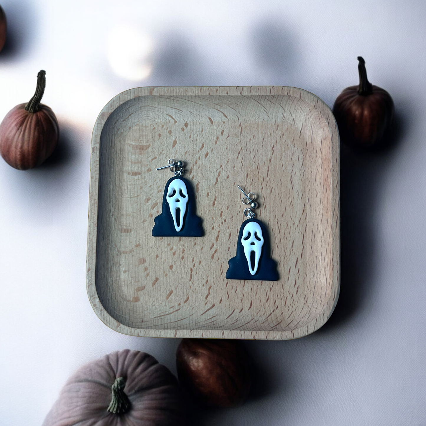 Black and white Ghostface-inspired horror earrings resting in a wooden tray, surrounded by pumpkins for a spooky, minimalist vibe. (