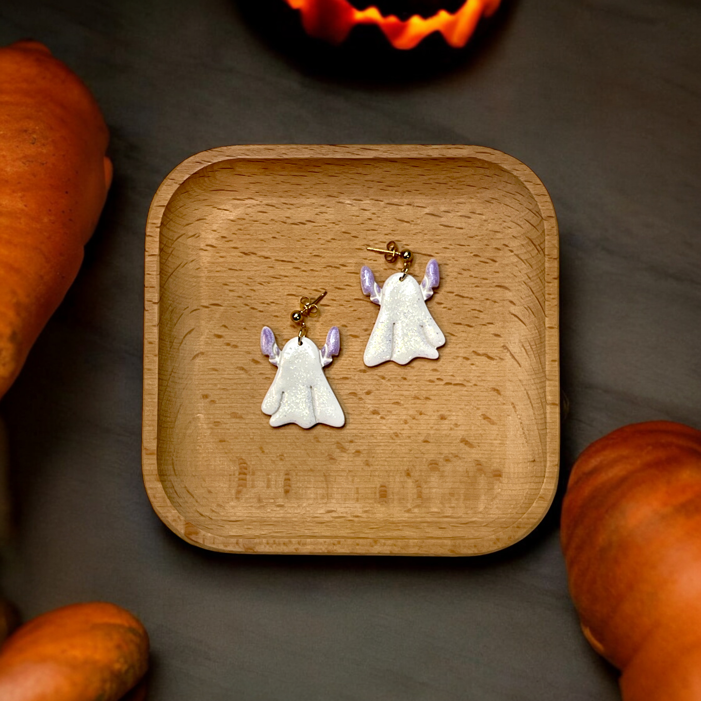 Pair of glittery ghost earrings with lavender accents placed on a wooden tray, surrounded by pumpkins and soft Halloween-themed lighting.