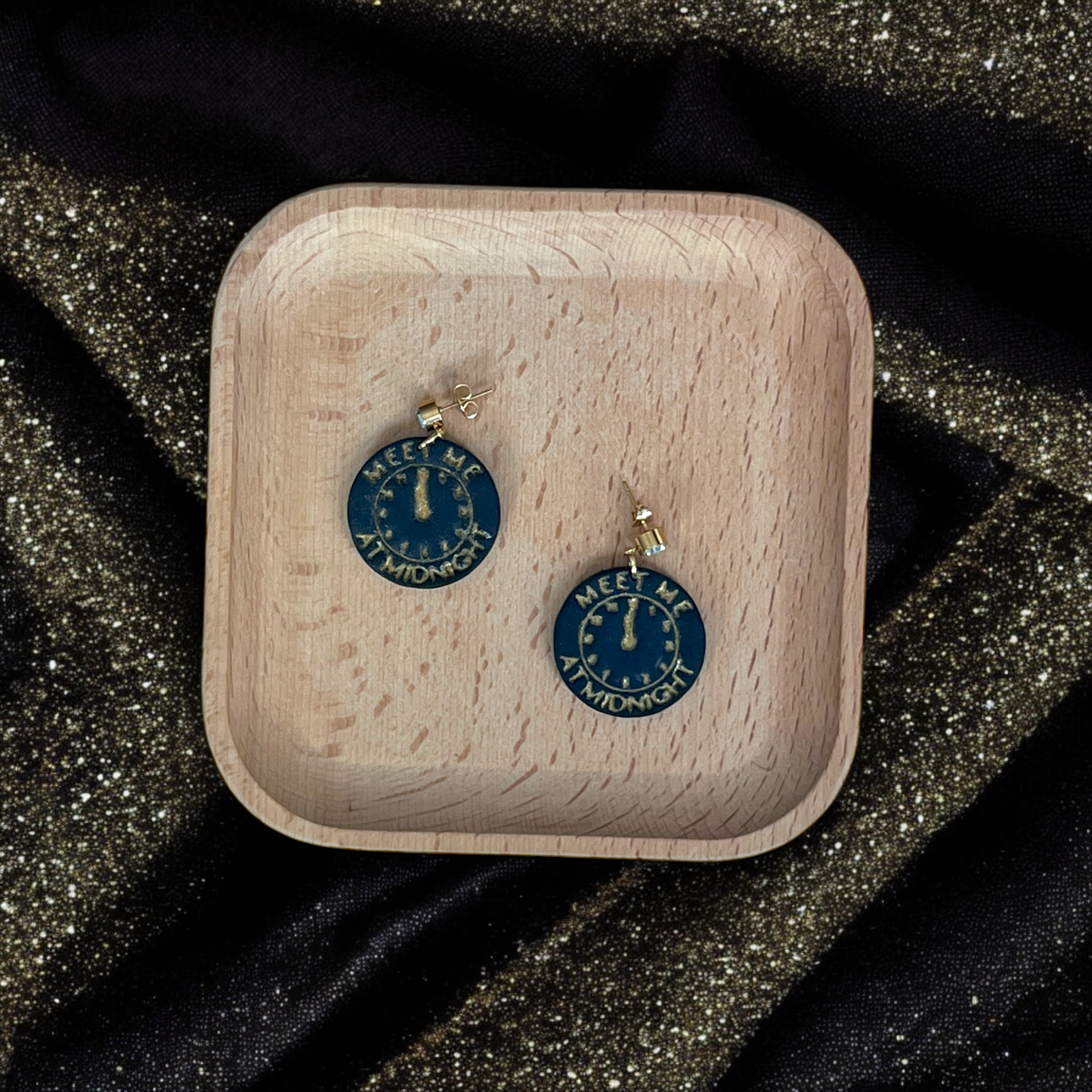 Circular earrings displaying a gold clock striking midnight with engraved text, placed on a wooden tray surrounded by shimmering black and gold fabric.