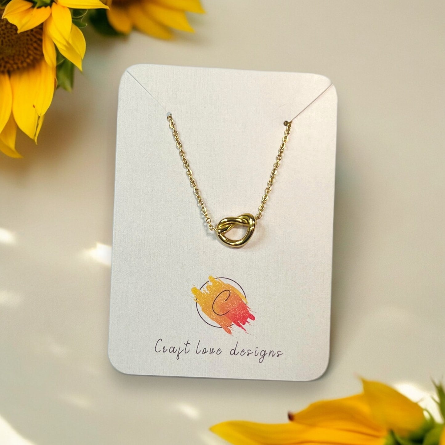 Delicate gold knot pendant necklace presented on a Craft Love Designs display card with sunflower accents.