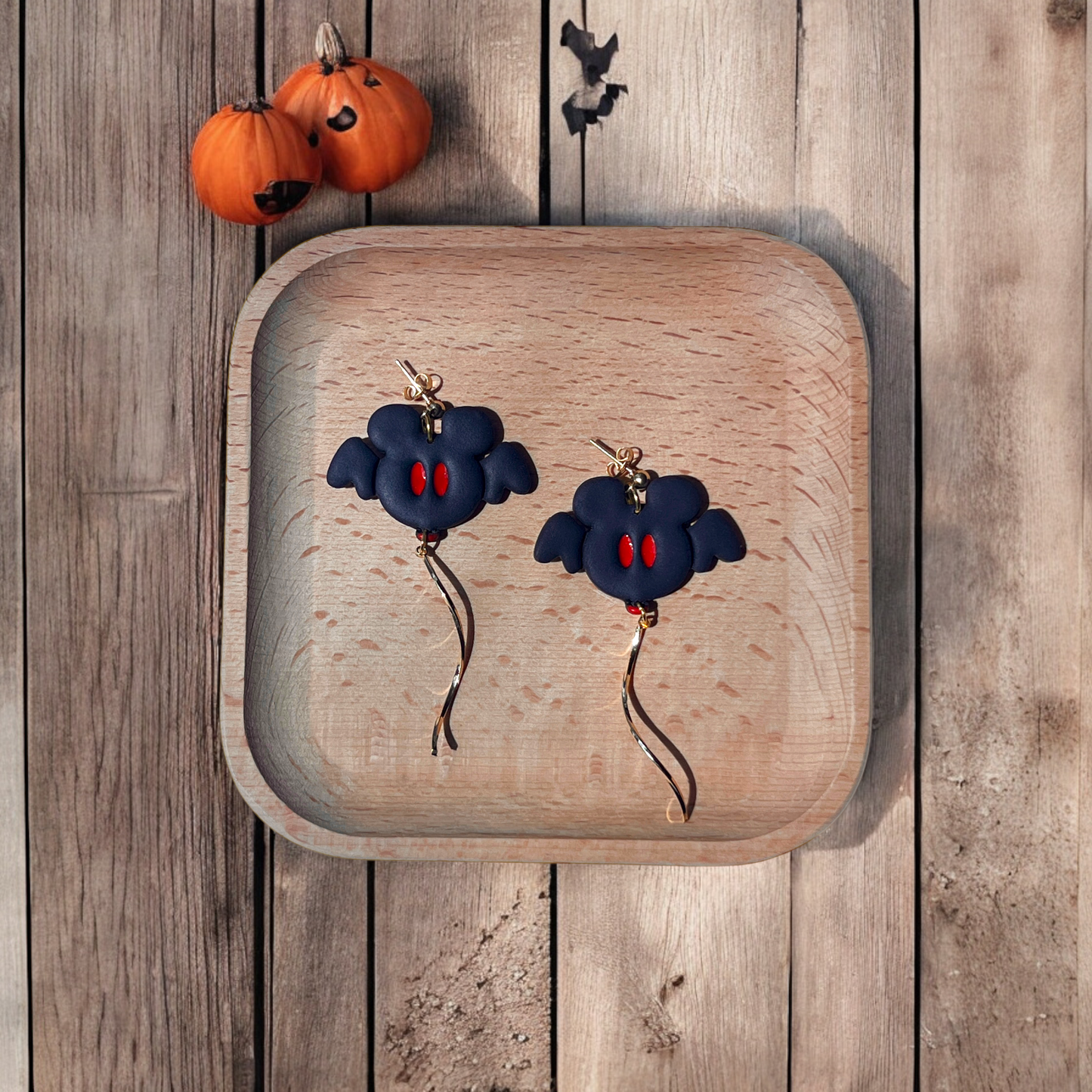 Polymer clay bat earrings with red eyes and gold dangle charms, set in a wooden tray with rustic autumn decorations.