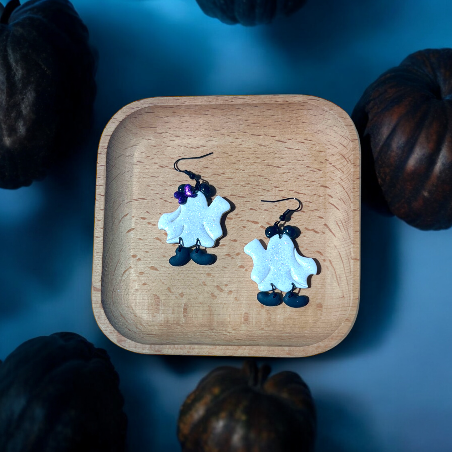 A pair of sparkly white ghost earrings with purple bow accents and black dangling shoes, displayed on a wooden tray with a festive Halloween setting.