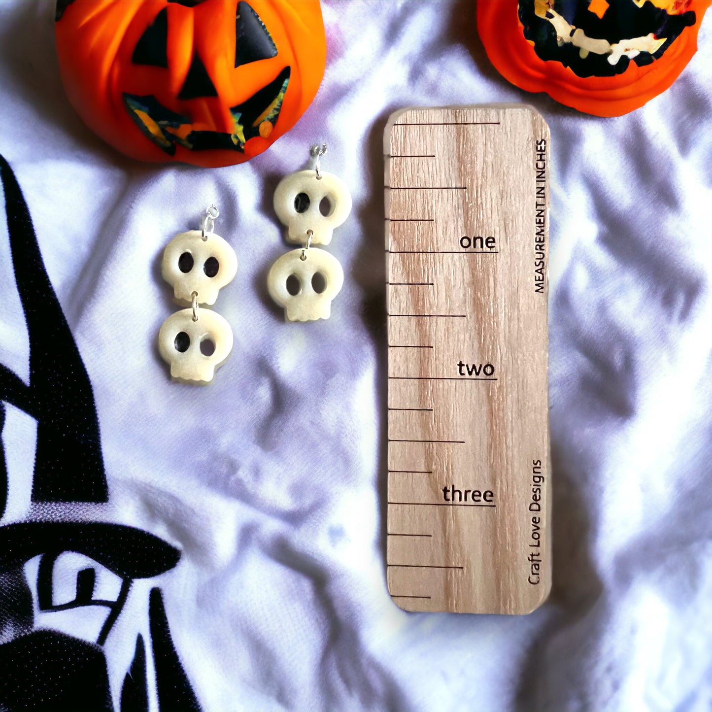 Pair of white glow-in-the-dark skull earrings displayed next to a wooden measurement scale and festive Halloween pumpkins.
