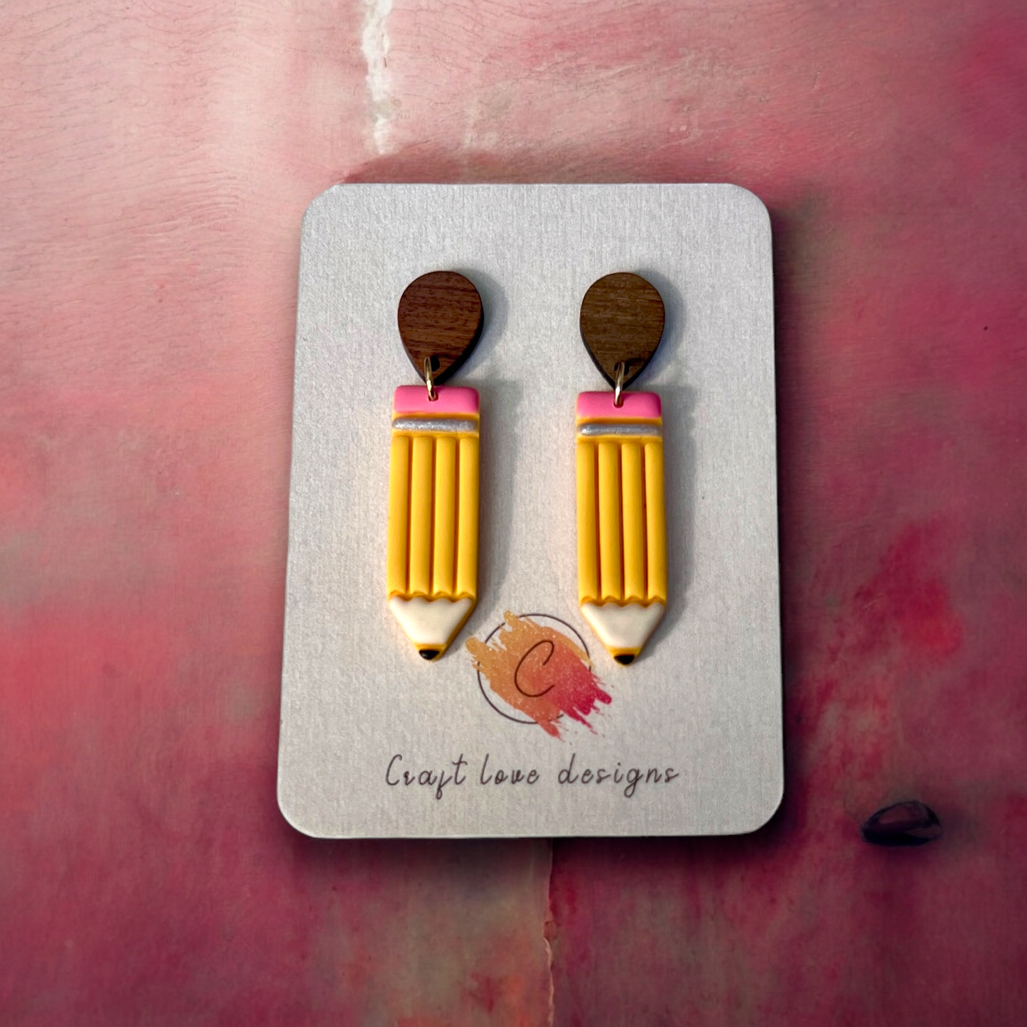 Whimsical handmade yellow pencil earrings with pink erasers, attached to wooden studs, displayed on a designer card over a soft pink background.