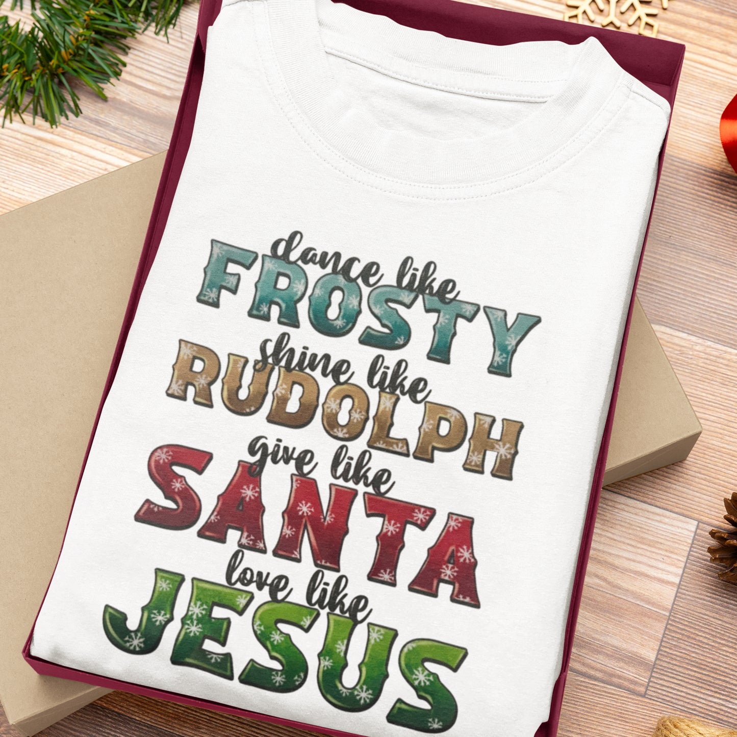 White Christmas-themed t-shirt folded inside a gift box, featuring festive text: "Dance like Frosty, Shine like Rudolph, Love like Jesus."