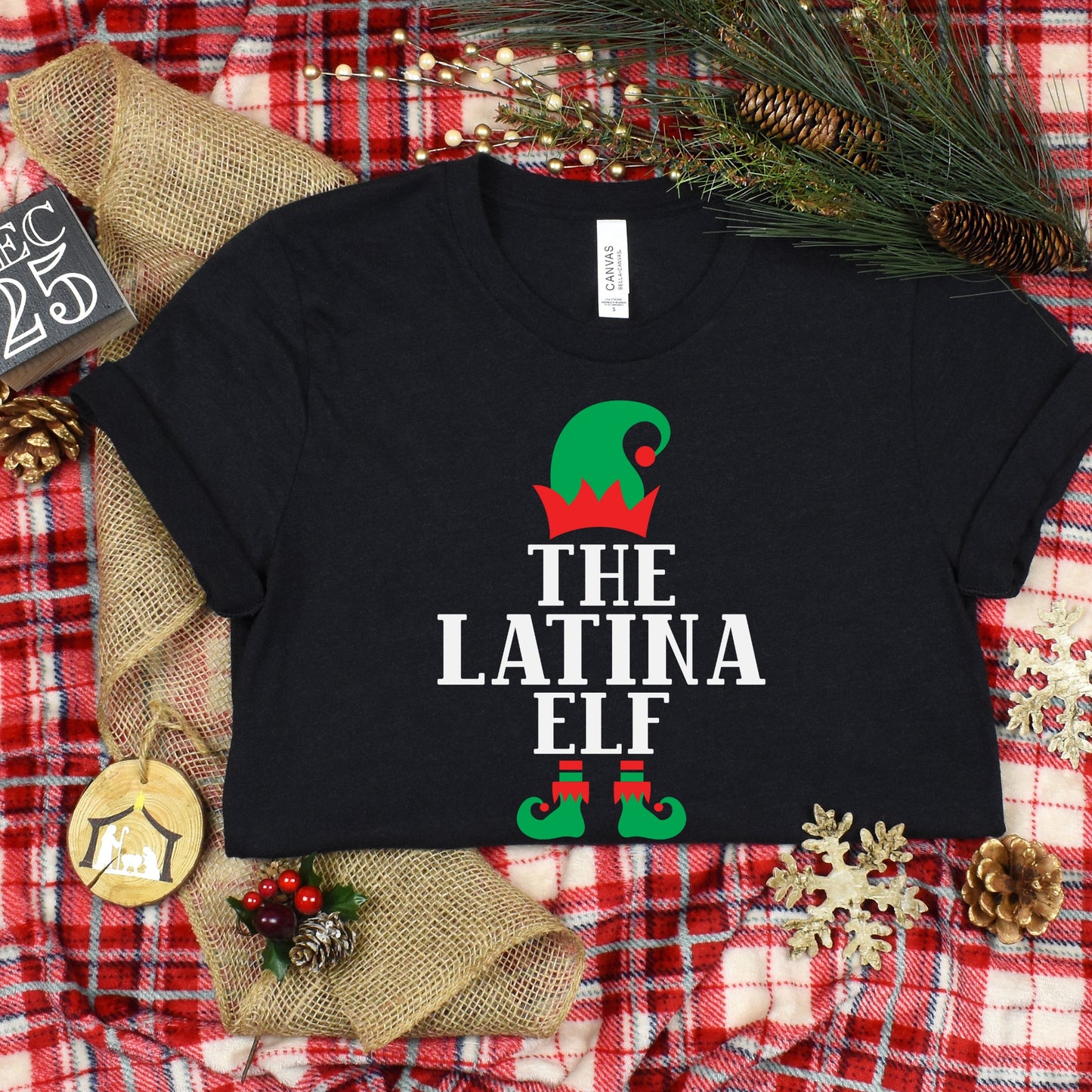 Flat lay of black "The Latina Elf" Christmas t-shirt with festive red, green, and white design, displayed on a plaid holiday background.