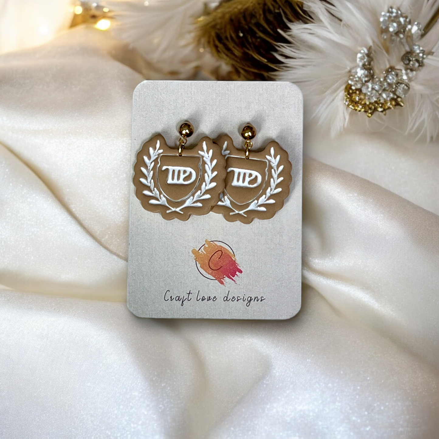 Beige dangle earrings featuring a laurel crest design displayed on a Craft Love Designs card, set on silky white fabric with soft accents.