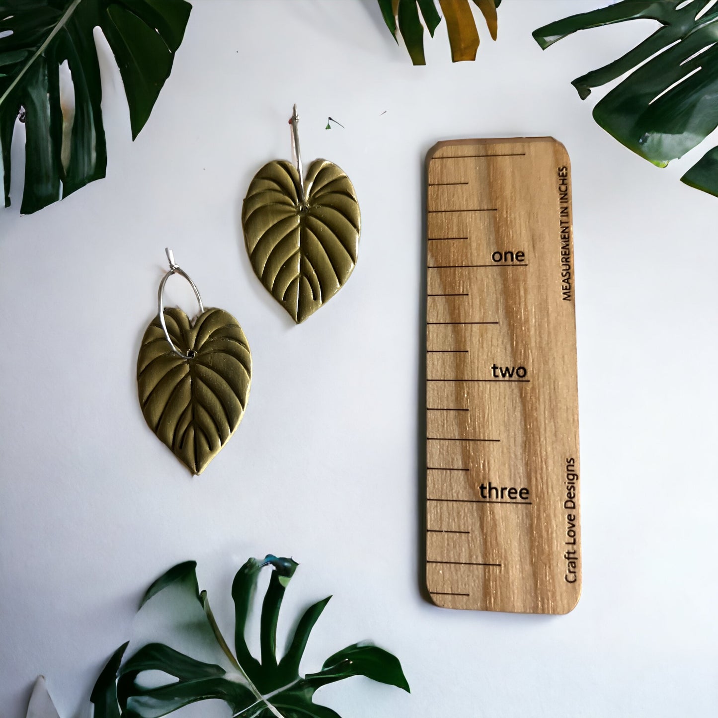 Pair of olive green philodendron leaf hoop dangle earrings displayed alongside a wooden measurement guide on a white background.